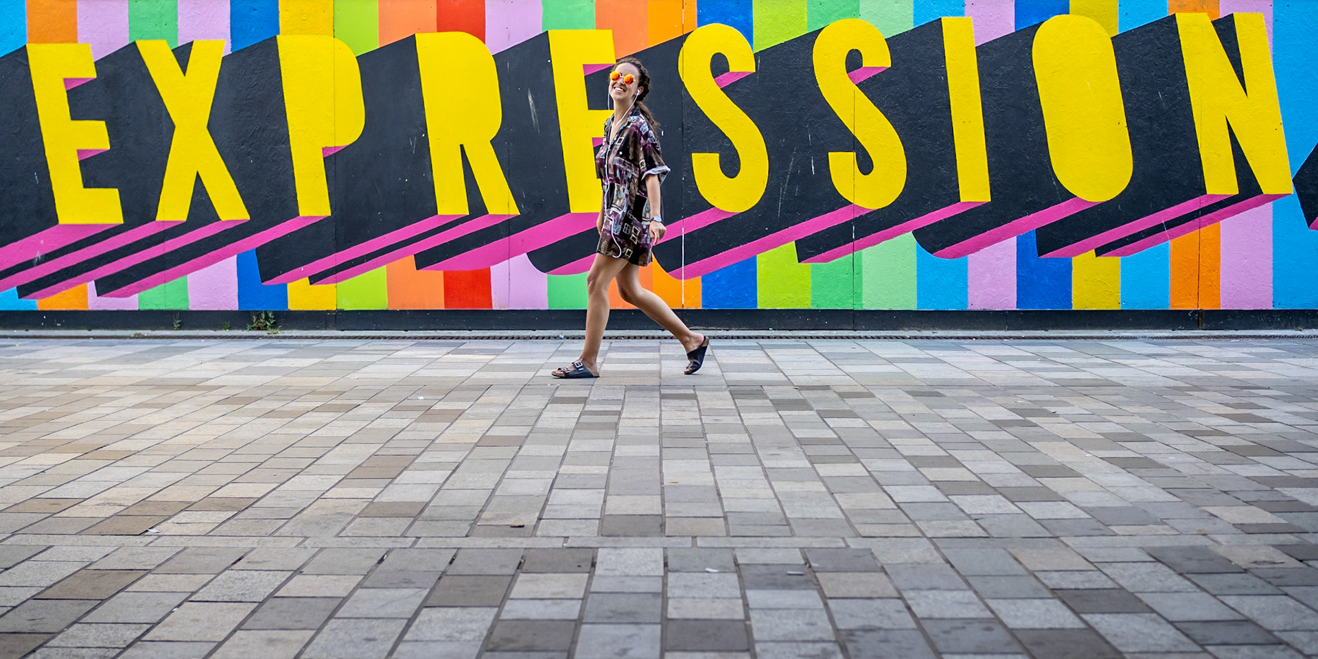 A carefree young woman wearing tinted sunglasses strolls casually in front of colourful hoarding outside Brighton's Dome Theatre