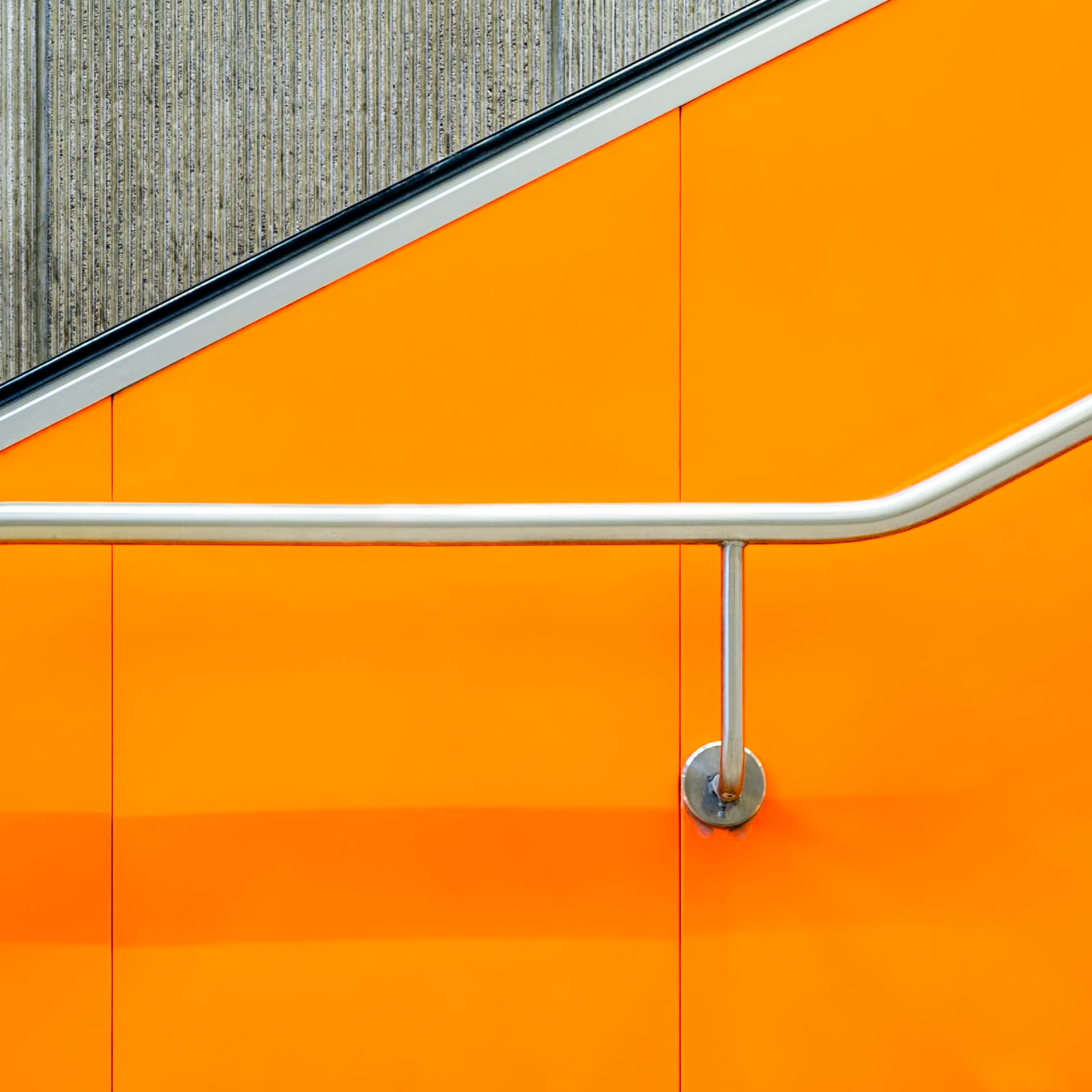 A chrome handrail in stark contrast to the bright orange panelling of an escalator, a stark contrast to the concrete wall behind