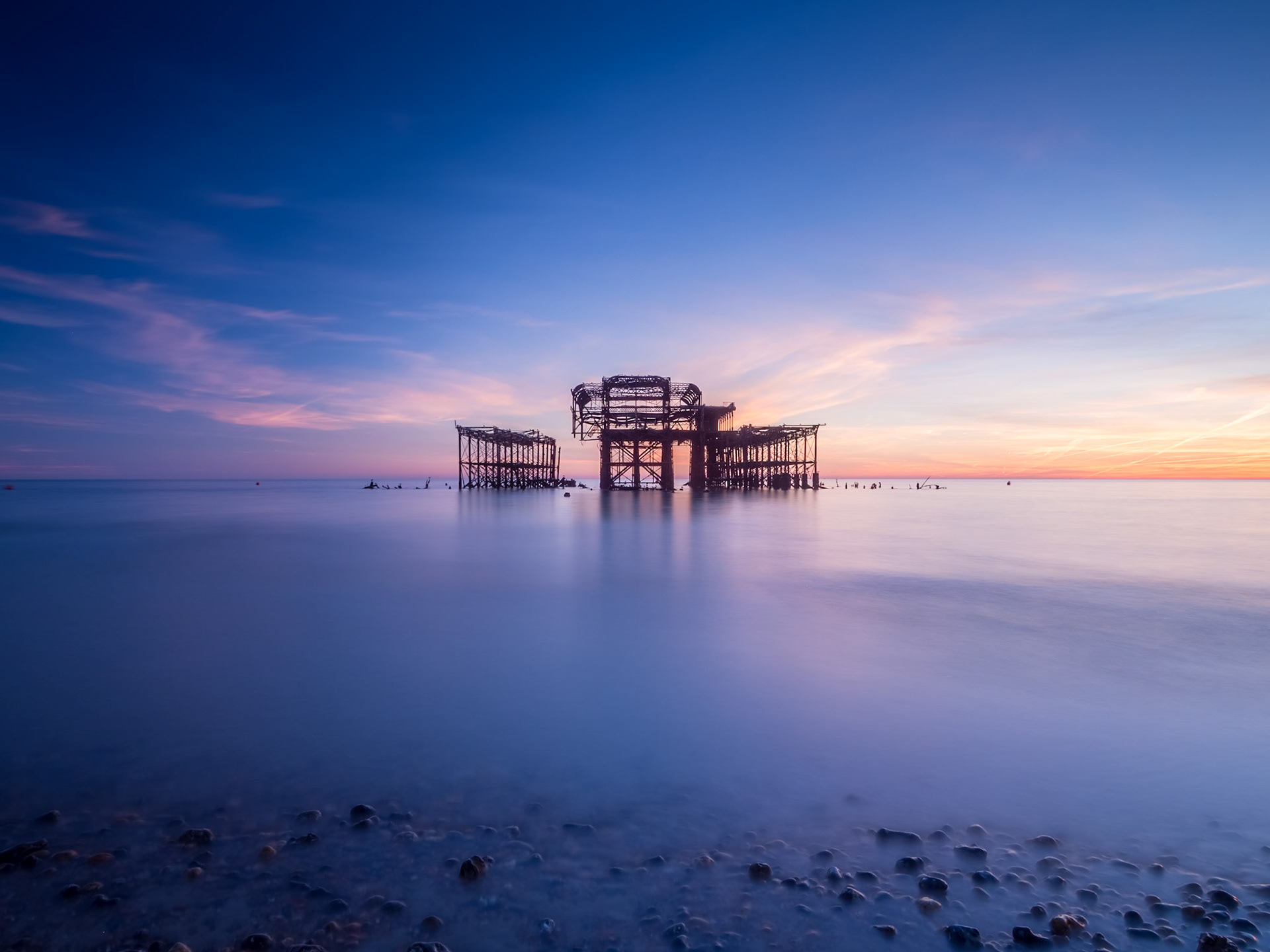 Pebbles emerge from a calm sea as the sun sets behind the derelict West Pier off Brighton seafront