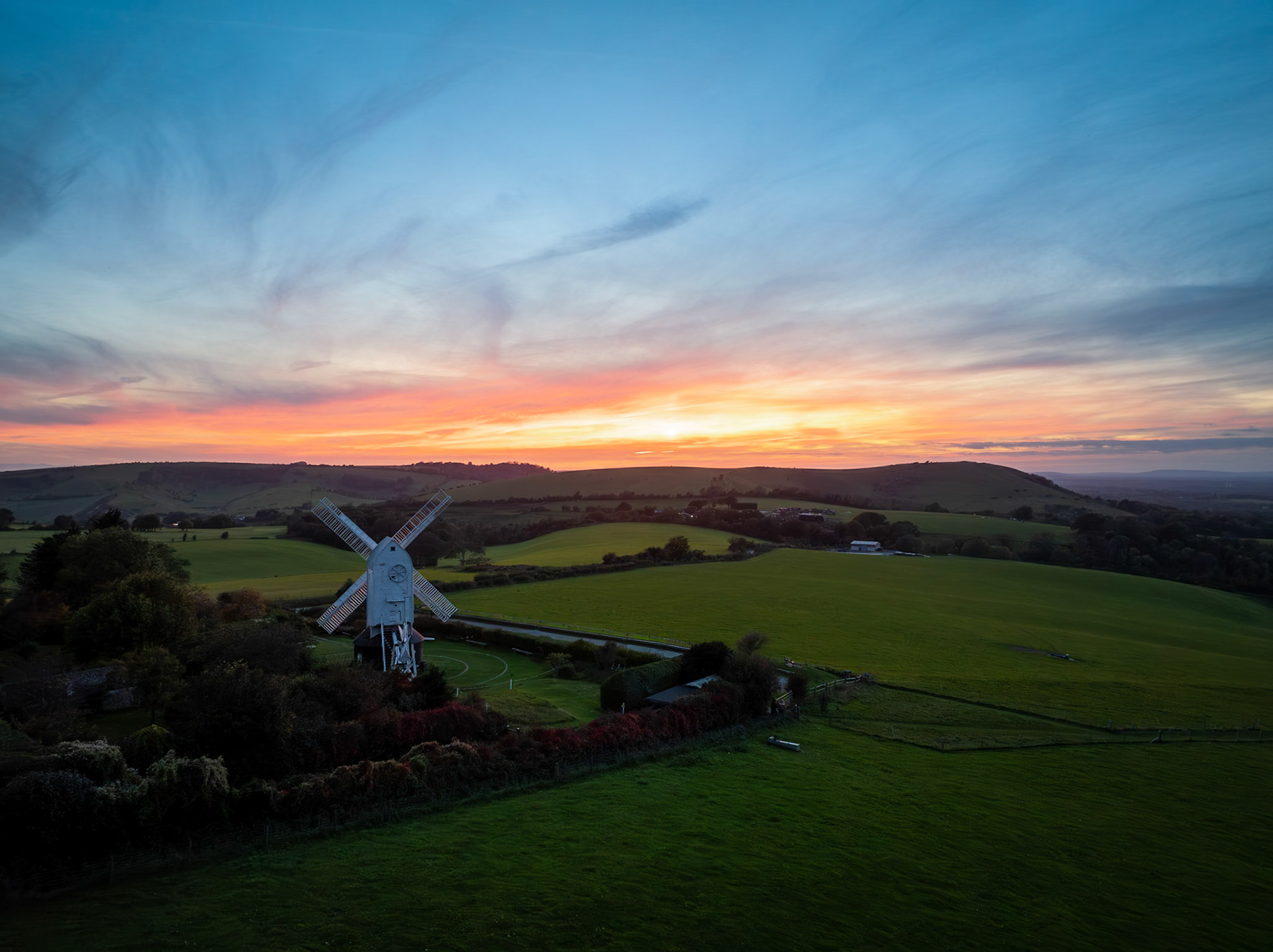 The vast South Downs landscape is bathed in the brilliant fire of the vibrant sunset, perfectly framing the iconic white sails of the solitary windmill.