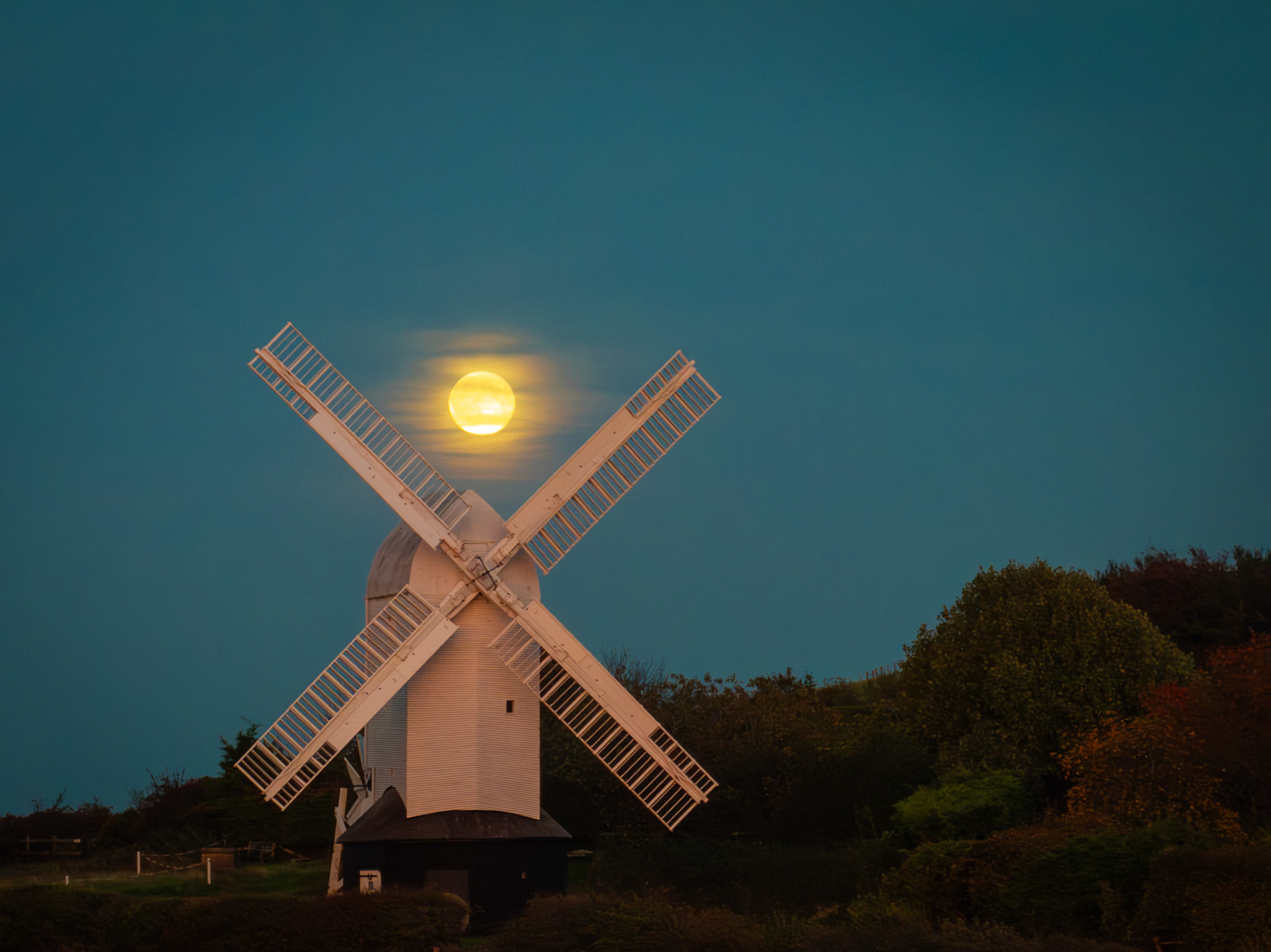 The enormous Harvest Moon casts a luminous, soft glow, its presence momentarily veiled by delicate wisps of cloud and starkly contrasted by the dark windmill sails on the South Downs.