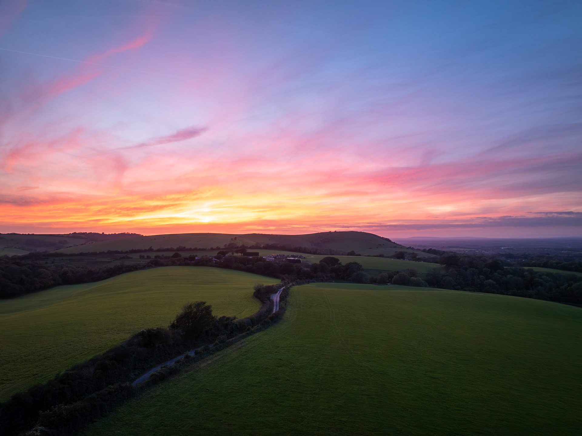 As dusk settles over the rolling hills, the last light of the vibrant sunset paints the sky in fire and pastel, bringing a peaceful silence to the South Downs.