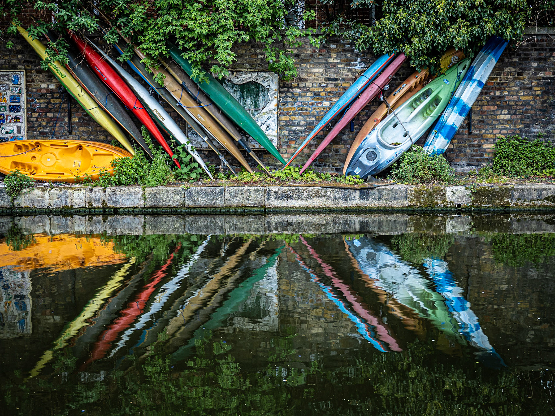 A collection of canoes reflect in the water as they rest against the banks of the Regents Canal in London
