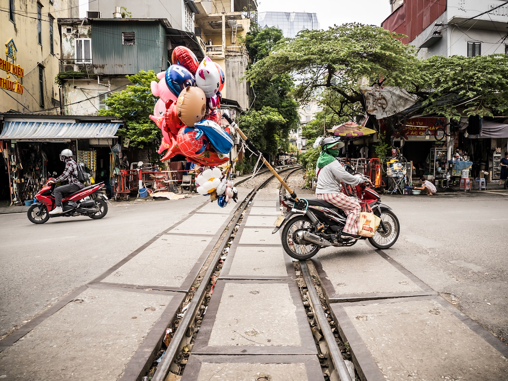 A woman guides her moped, laden with a magnificent bouquet of colourful balloons, across the railway tracks of Hanoi, transforming an everyday commute into a moment of pure, unexpected buoyancy