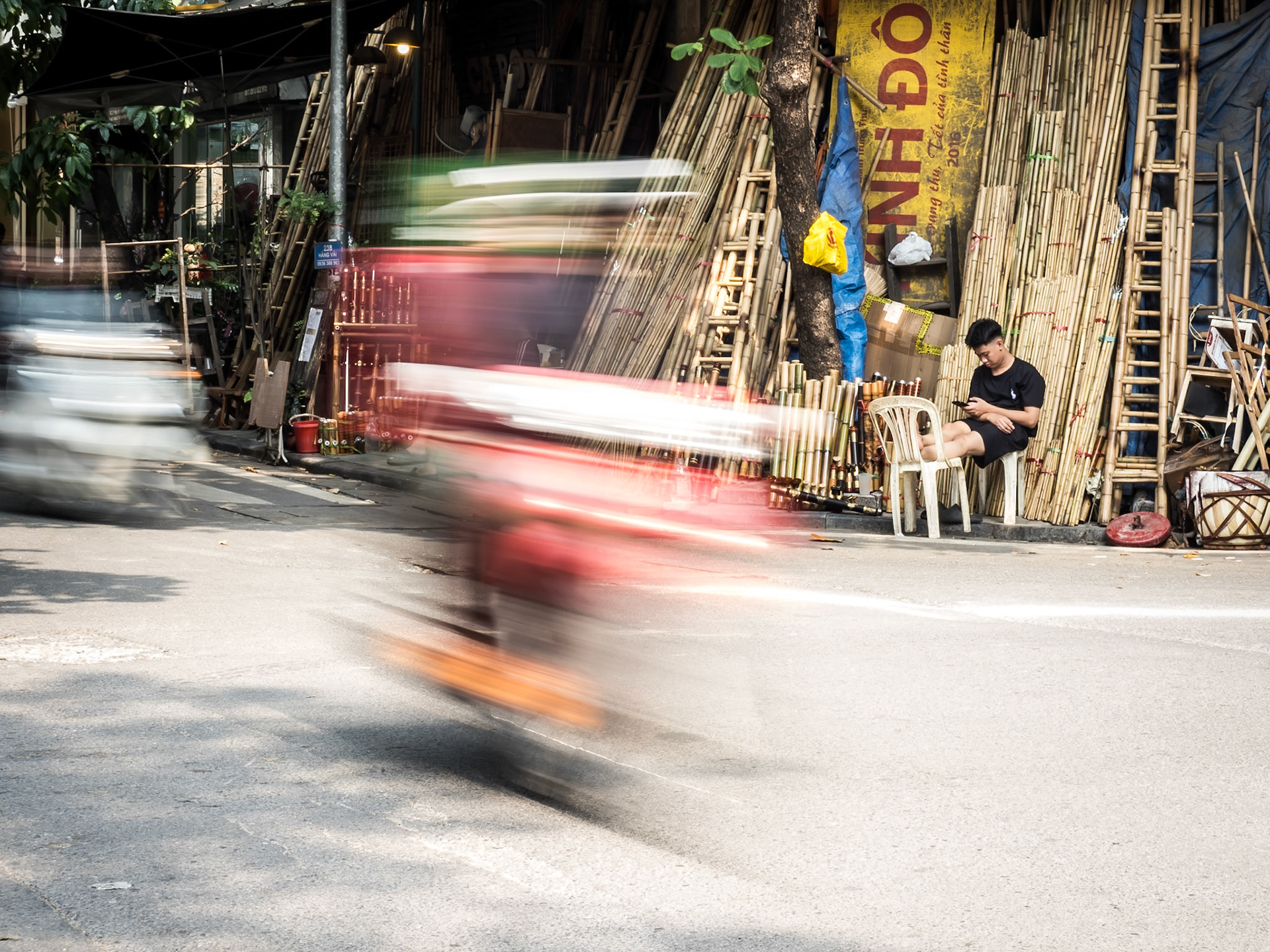 A moped blurs past on a Hanoi street, a flash of city speed that stands out against the still figure of a man chilling with his phone, backed by a wall of bamboo poles