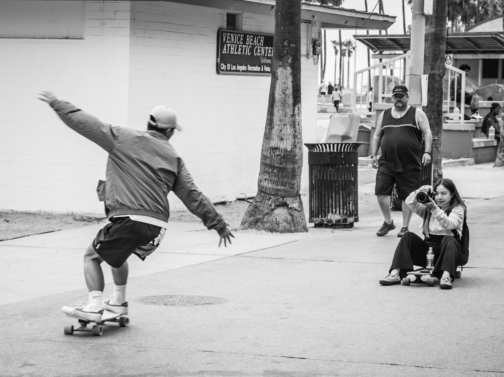 A girl captures her partner performing a skateboarding trick on Venice beachfront