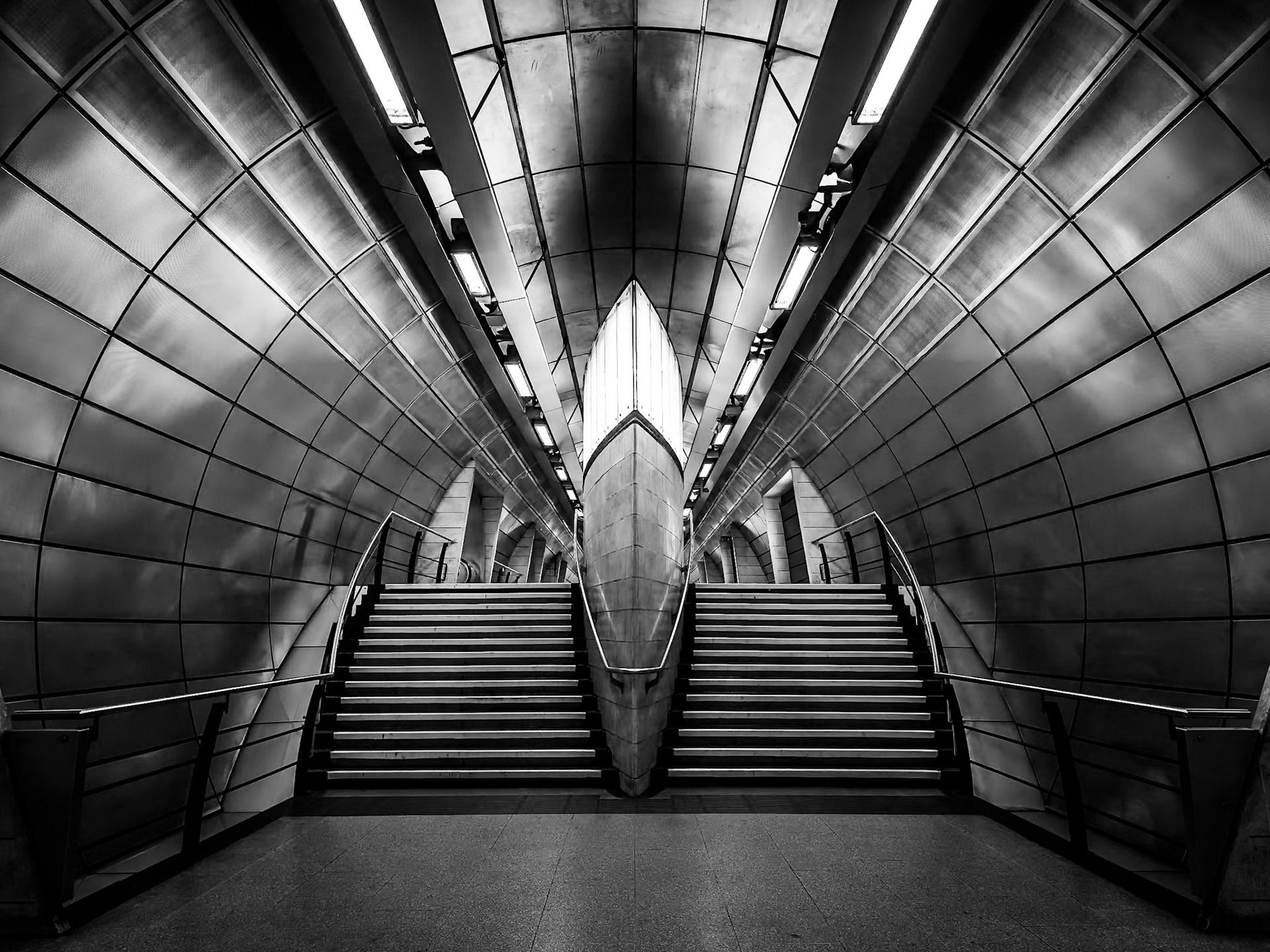 The intense symmetry and repetition of the metallic tunnel walls and steps create a powerful, stark form, drawing the eye deep into this modern, monochrome underground space.