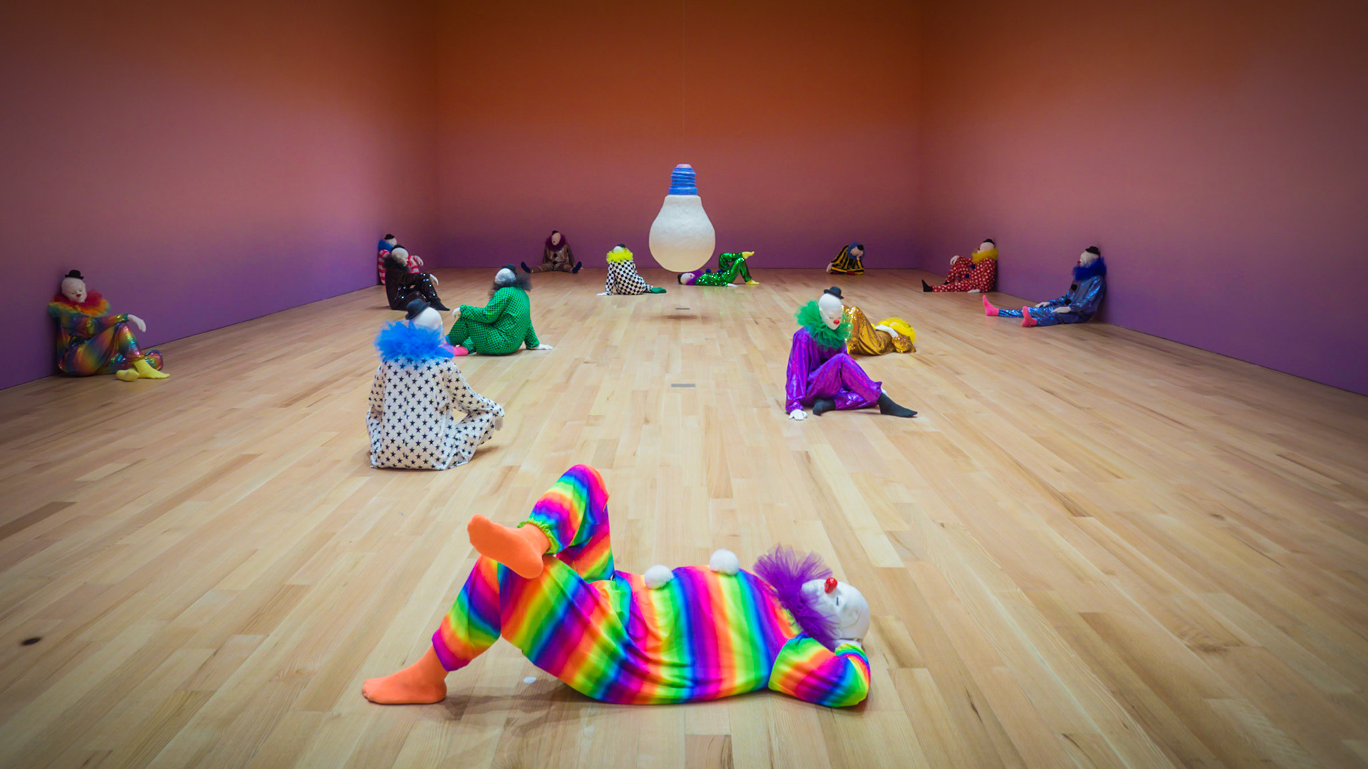 A contemporary art installation of clowns surrounding a light bulb