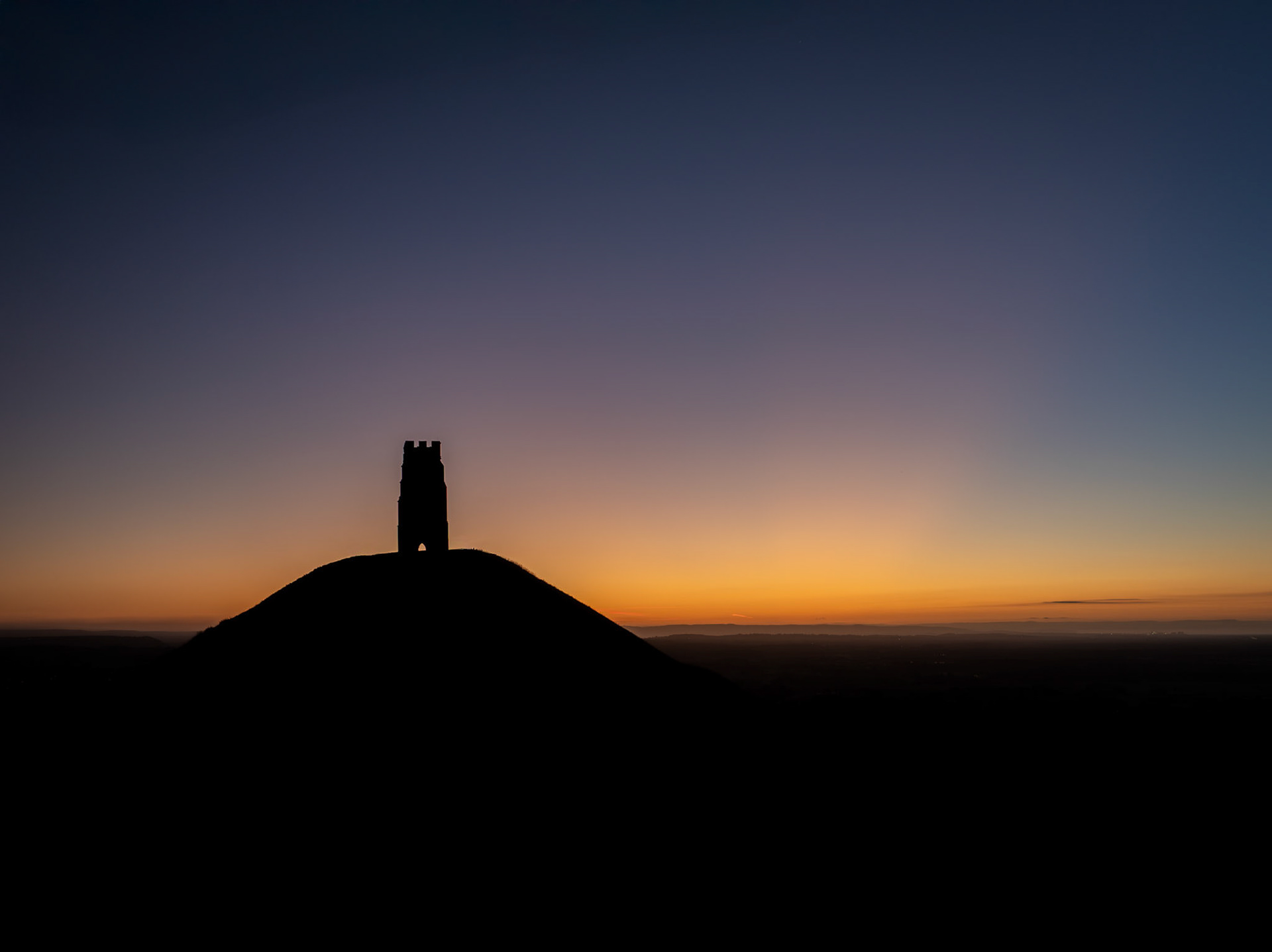 The vibrant, cloudless sunset creates a spectacular blaze of colour, backlighting Glastonbury Tor in an ethereal, almost mythical glow on the horizon.