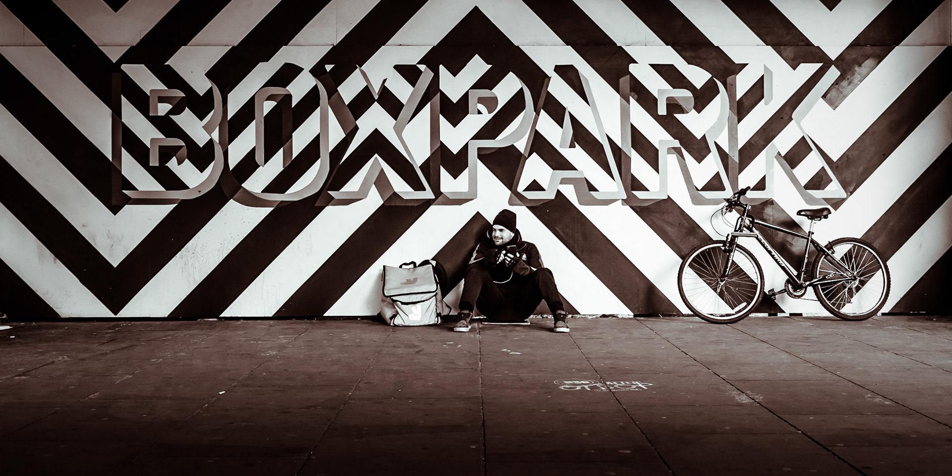 A fast food delivery worker chuckles as he rests between jobs in front of a contemporary sign in Shoreditch High Street