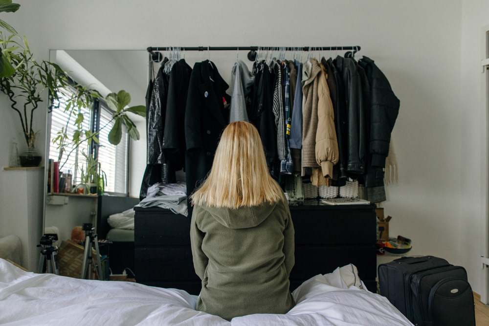 Blonde Frau auf Bett sitzend vor Kleiderschrank, Porträt von Oxana Guryanova.