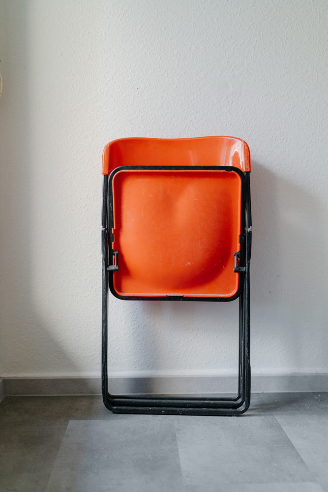 Orangener Klappstuhl an weißer Wand im Displaced-Projekt von Dokumentarfotografin Oxana Guryanova