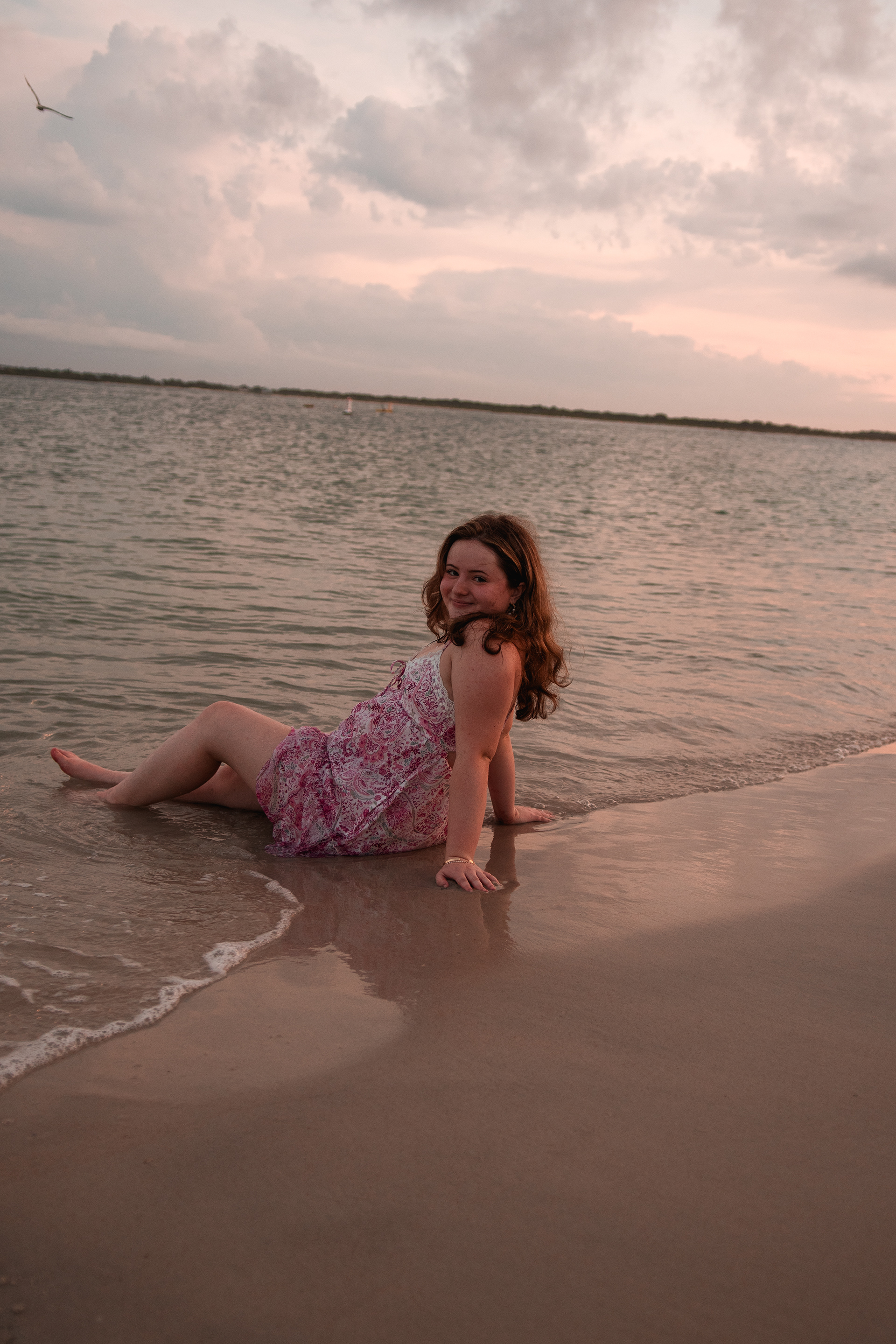 Senior portraits at Ponce Inlet, Nelly sitting in the wet sand at the shoreline in pink floral dress during golden sunset.