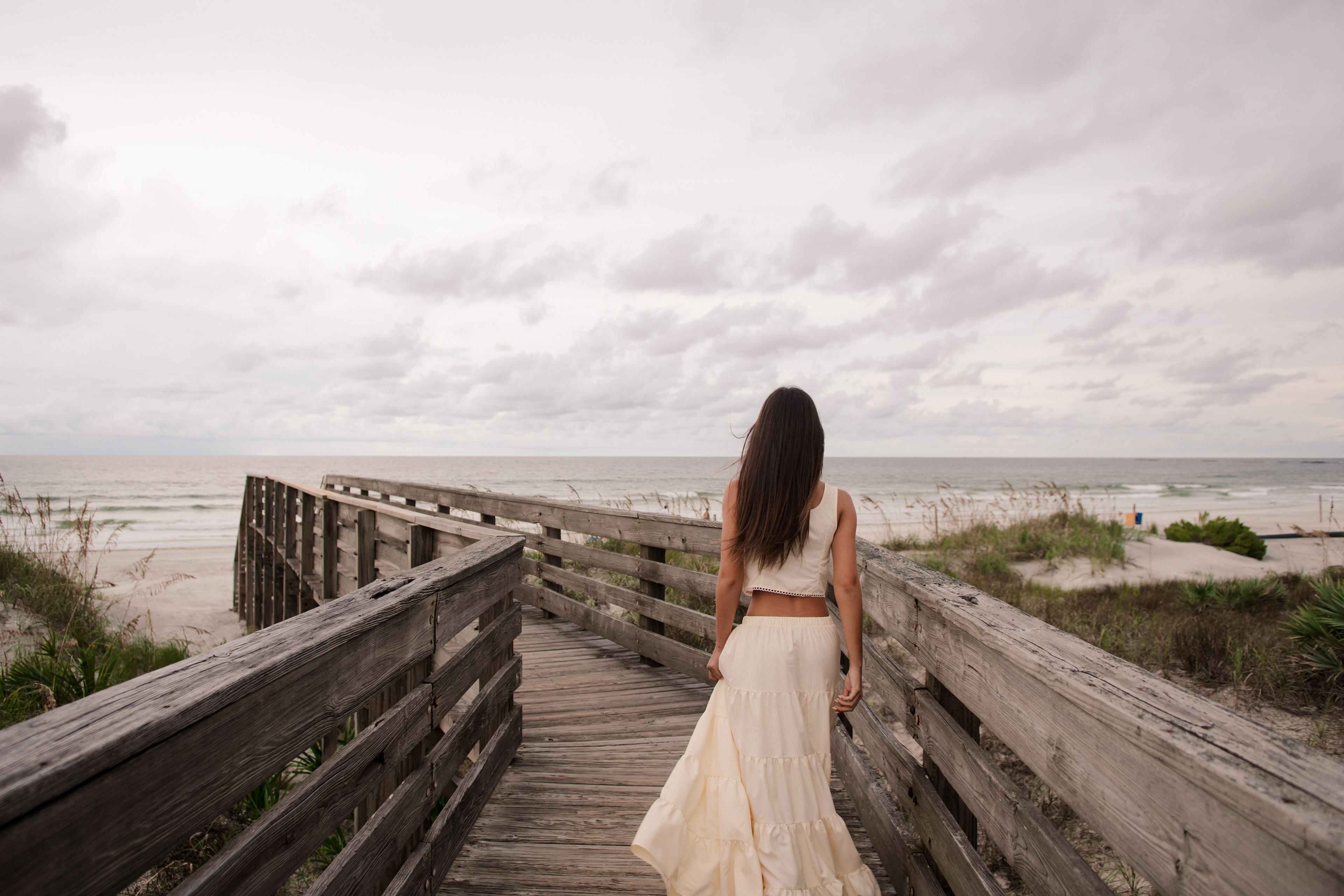 Senior portraits at Ponce Inlet Beach boardwalk, Mia walking toward the ocean in a cream skirt, candid moment.