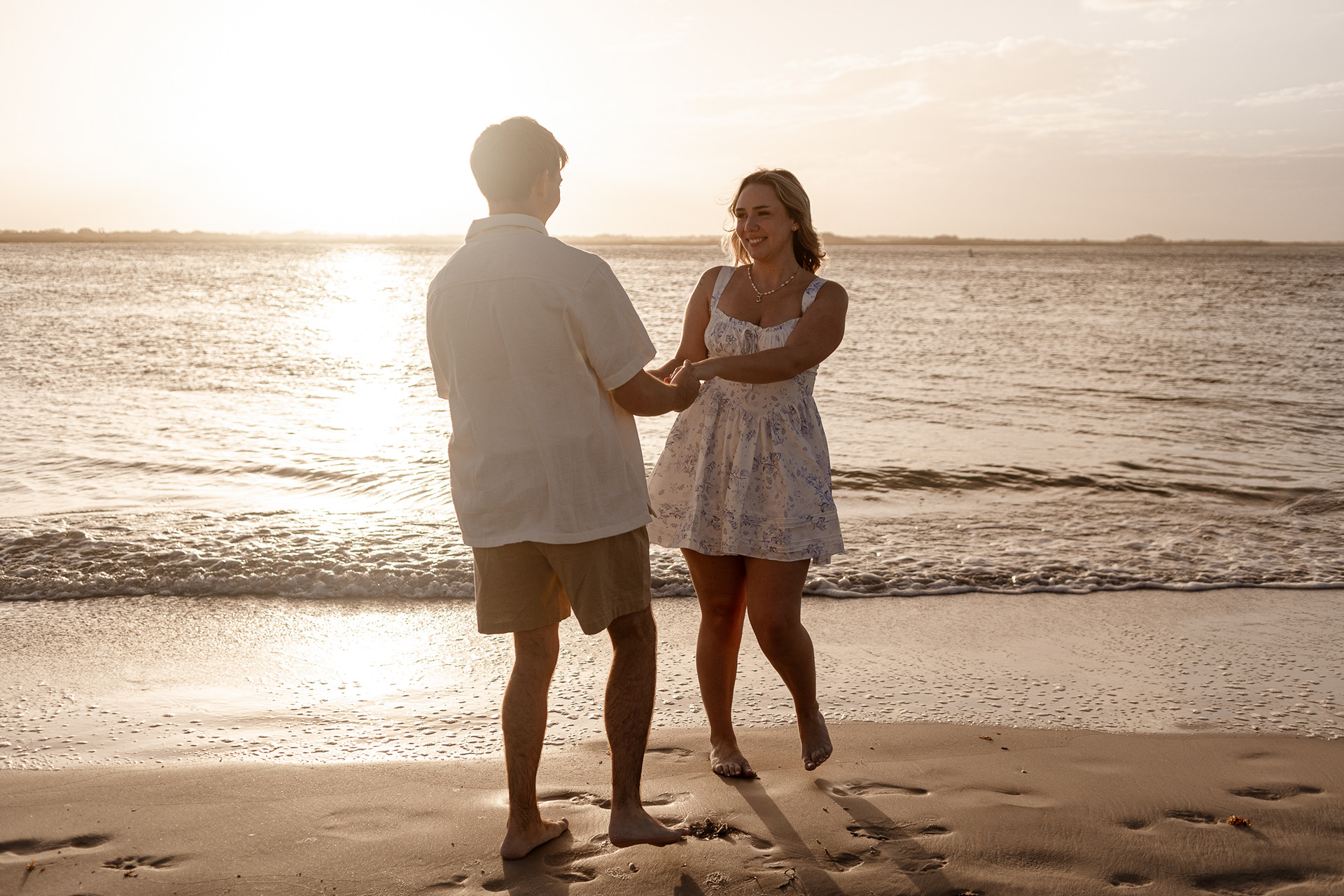 Sunset engagement photo in New Smyrna Beach, couple holding hands in shallow water with warm glow.