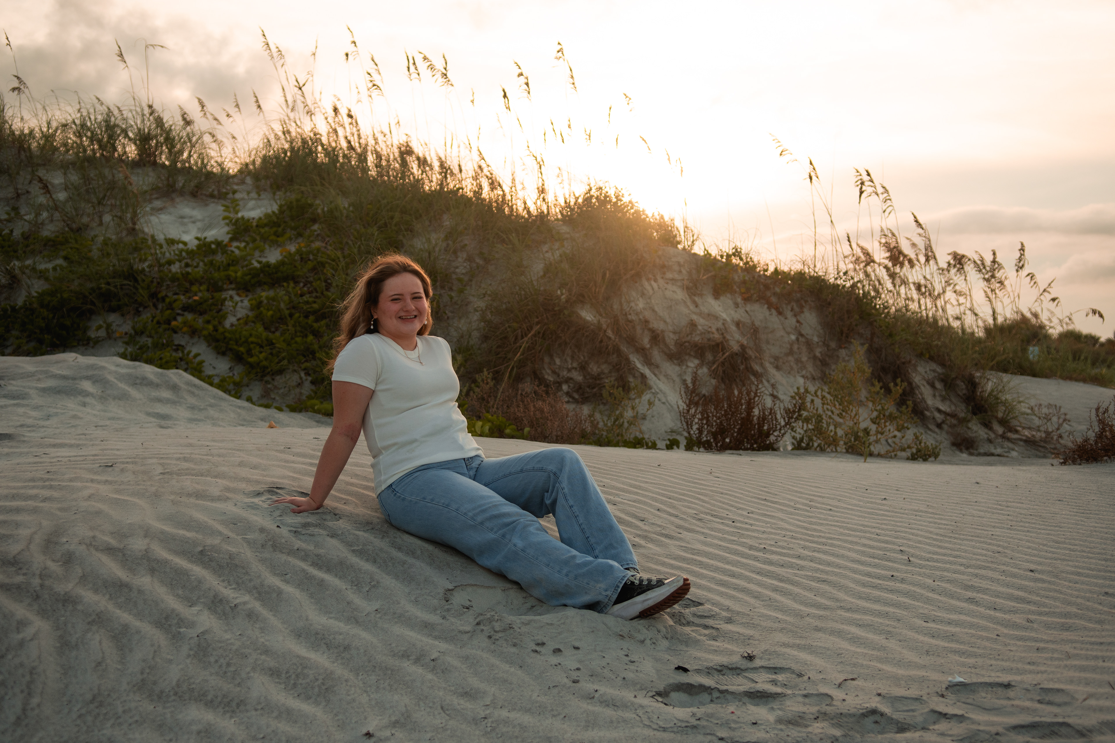 Senior photos in the dunes near Daytona Beach, Nelly sitting on the sand in jeans and white top with sea oats overhead.
