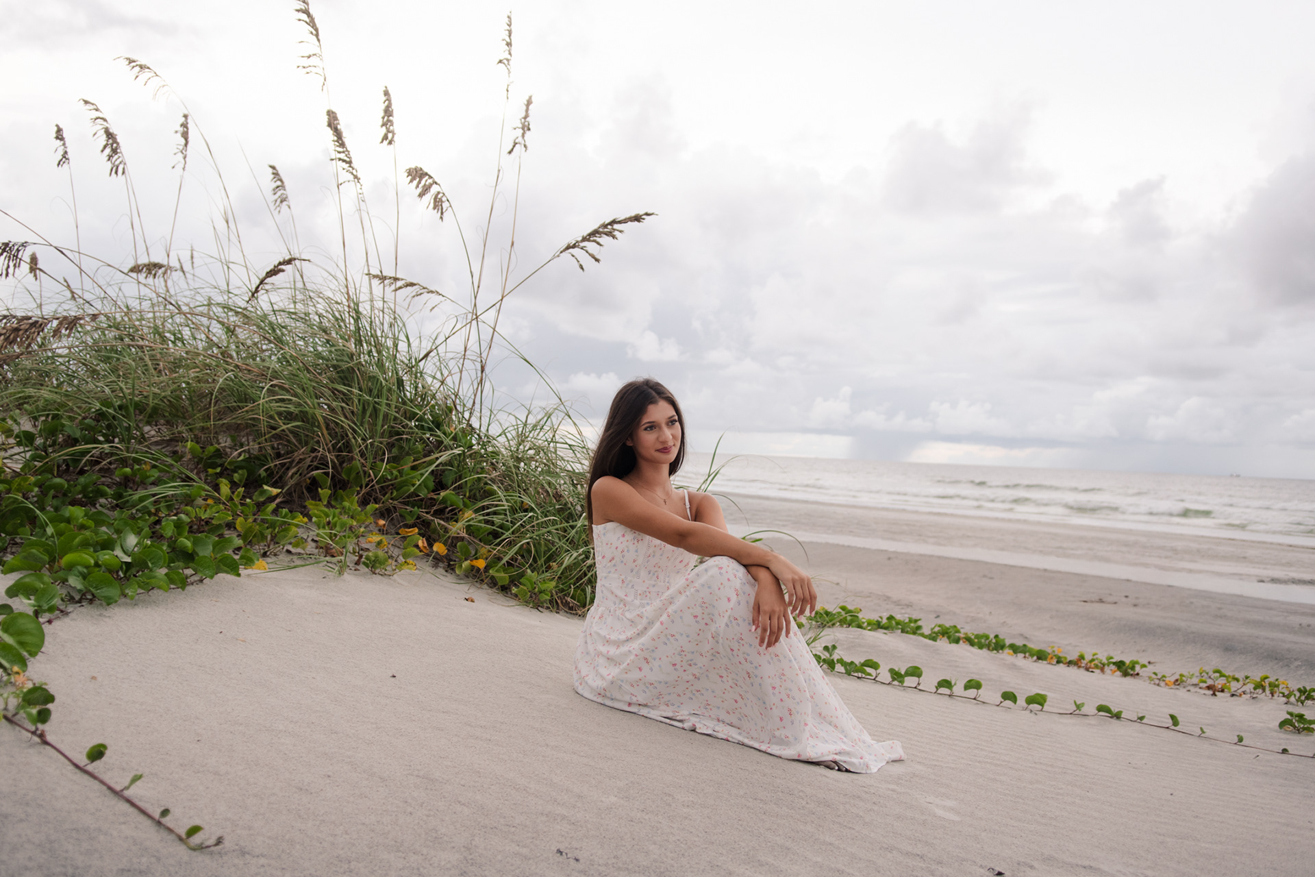 Senior photos in Ponce Inlet FL, Mia sitting in a white floral dress near the dunes with the beach in the background.