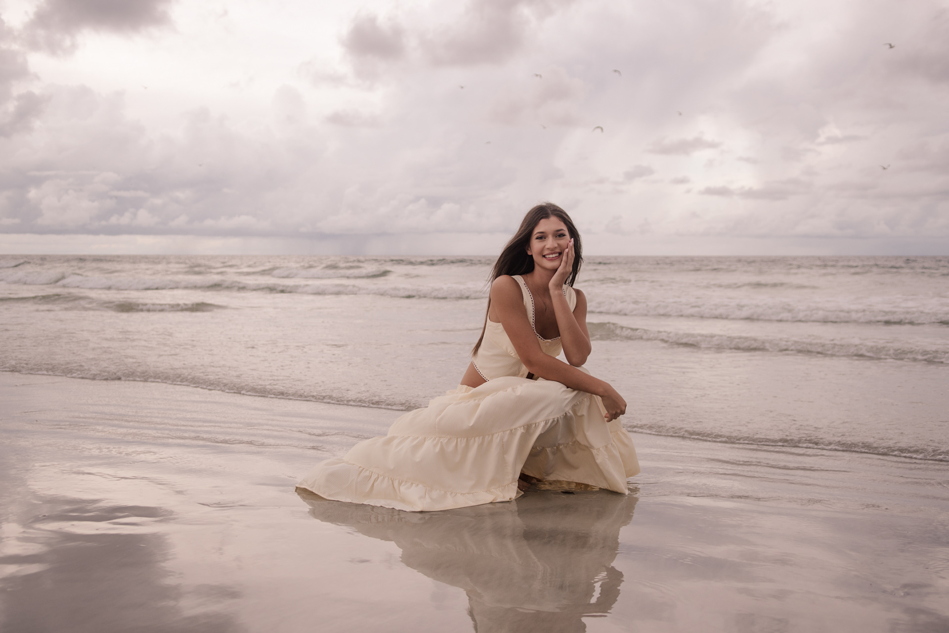 Senior portraits at Ponce Inlet Beach, Mia sitting at the shoreline in a cream dress with soft reflection in the water.