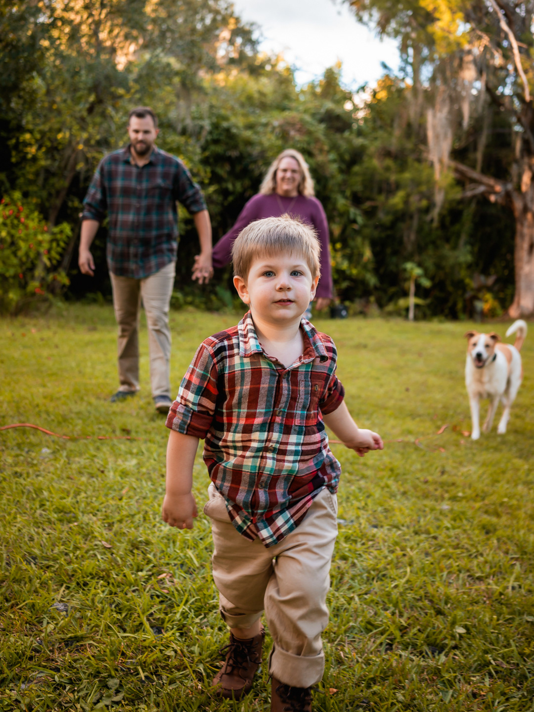 Playful family portrait in New Smyrna Beach, young boy running toward the camera with parents and dog in the background.