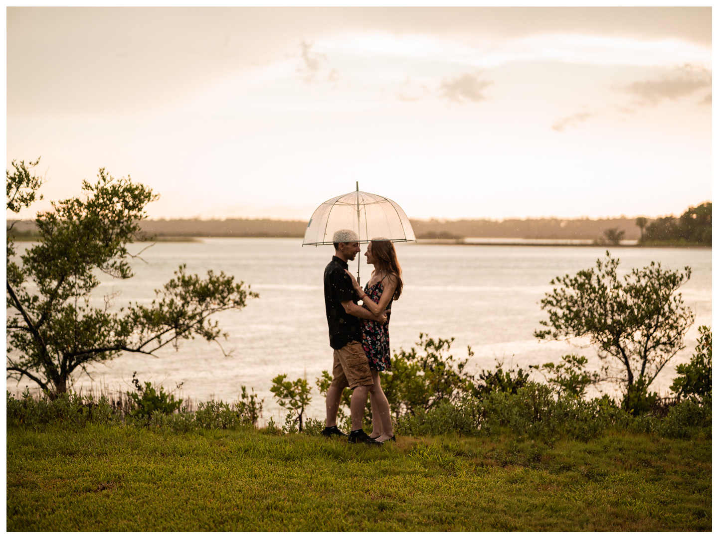 Wide portrait of a couple embracing beneath an umbrella on the grassy waterfront at Washington Oaks Gardens.