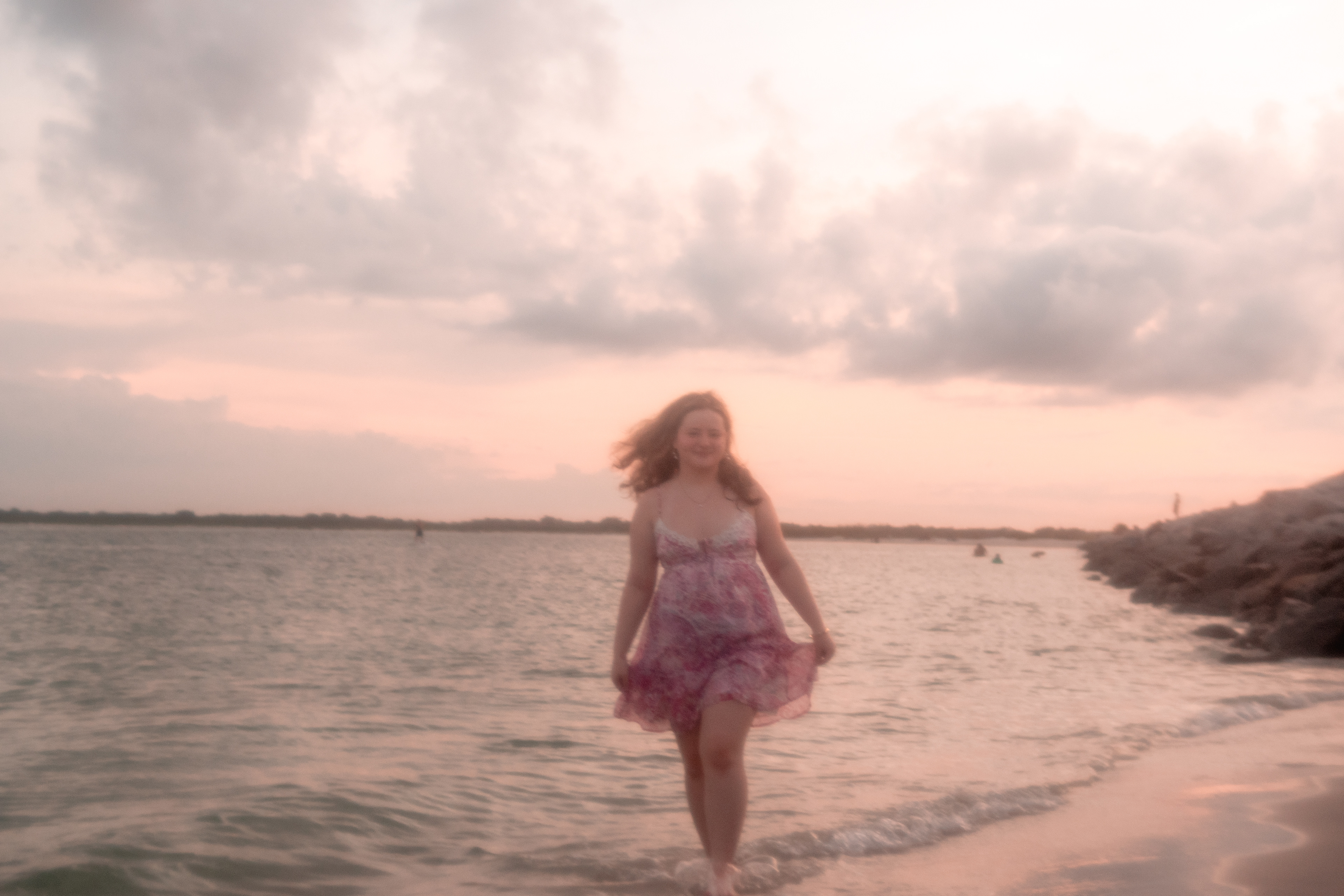Senior photo session at Ponce Inlet, Nelly seated on the sand in a pink dress with ocean waves rolling in.