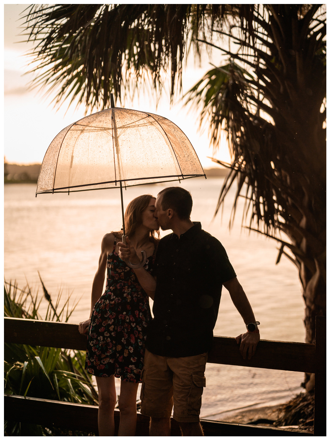 Couple kissing beneath a clear umbrella by a wooden railing overlooking the Matanzas River.