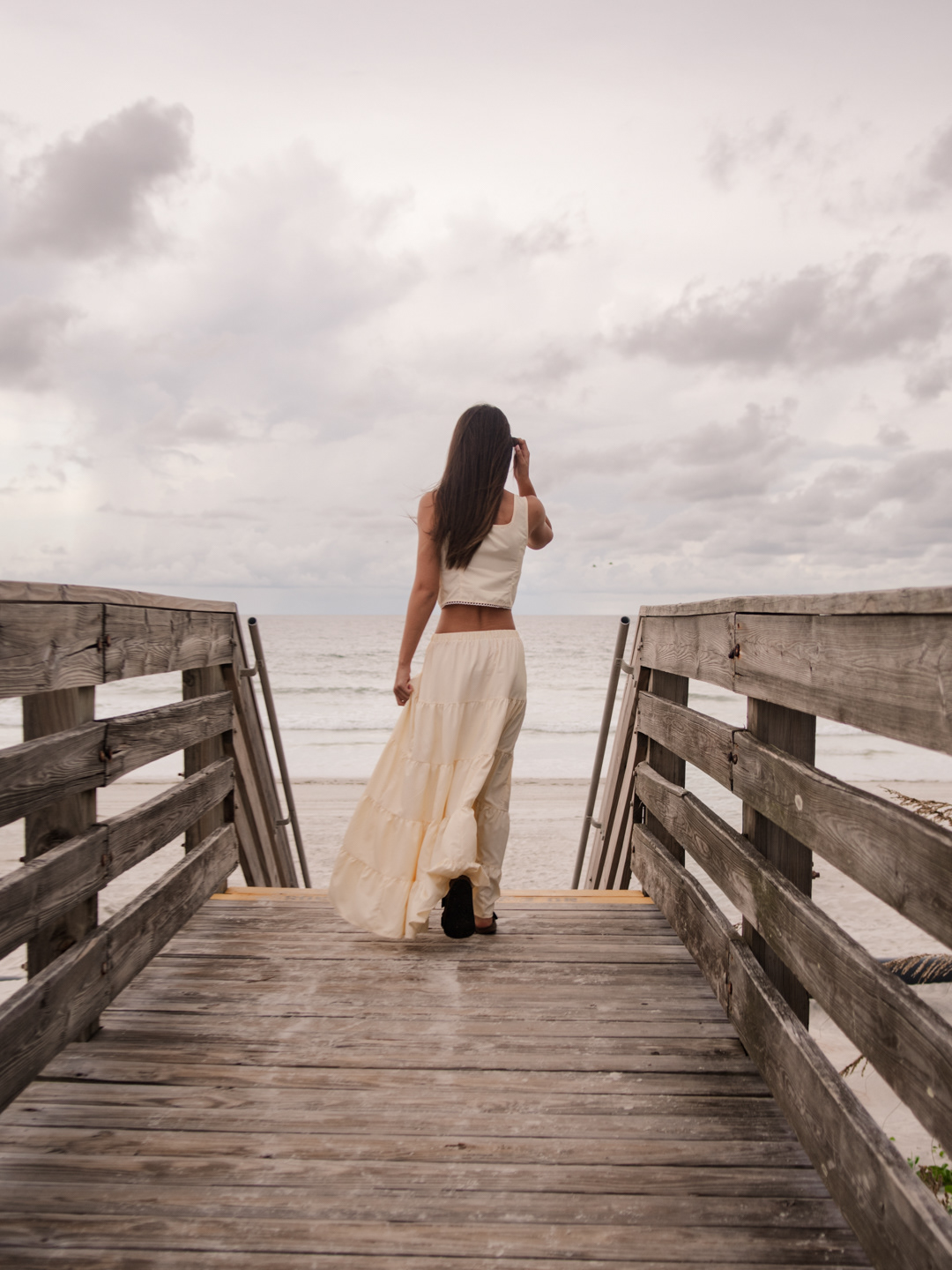 Senior photo session in Ponce Inlet FL, Mia walking on the wooden boardwalk in a cream maxi skirt with the ocean view ahead.
