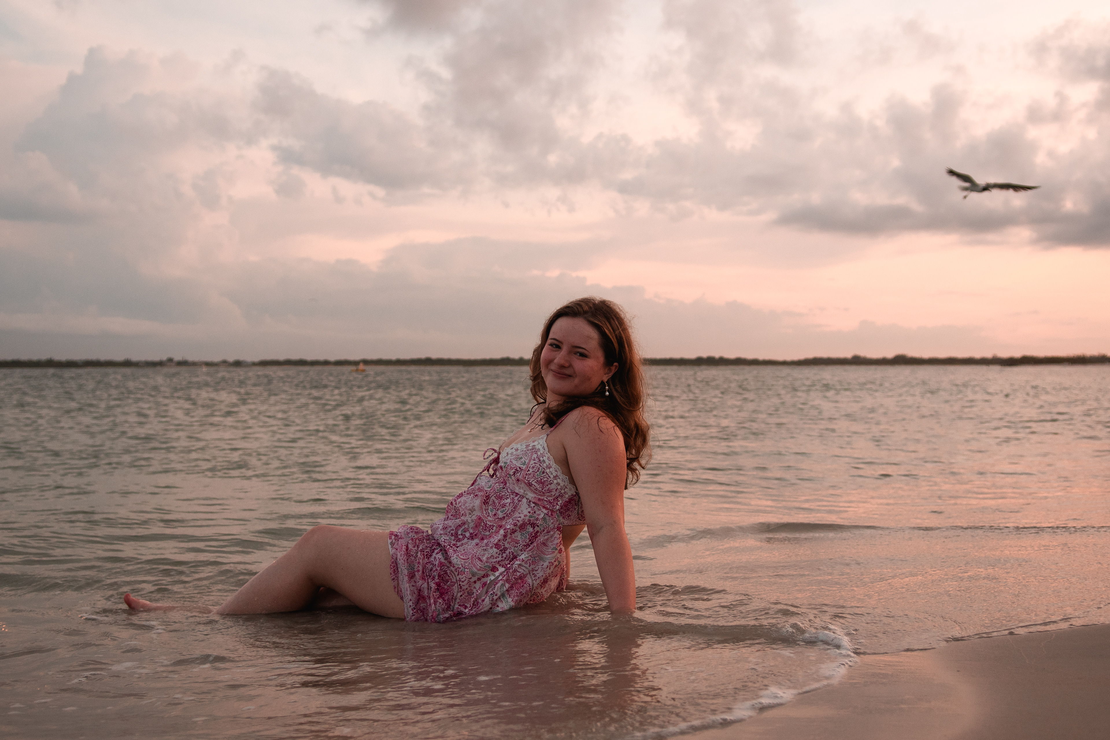 Daytona Beach area senior photos, Nelly smiling while seated at the water’s edge in pink dress, ocean glow behind her.
