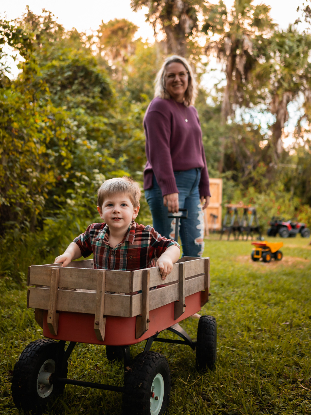 Little boy riding in a red wagon pulled by his mom during playful family photo session in New Smyrna Beach backyard.