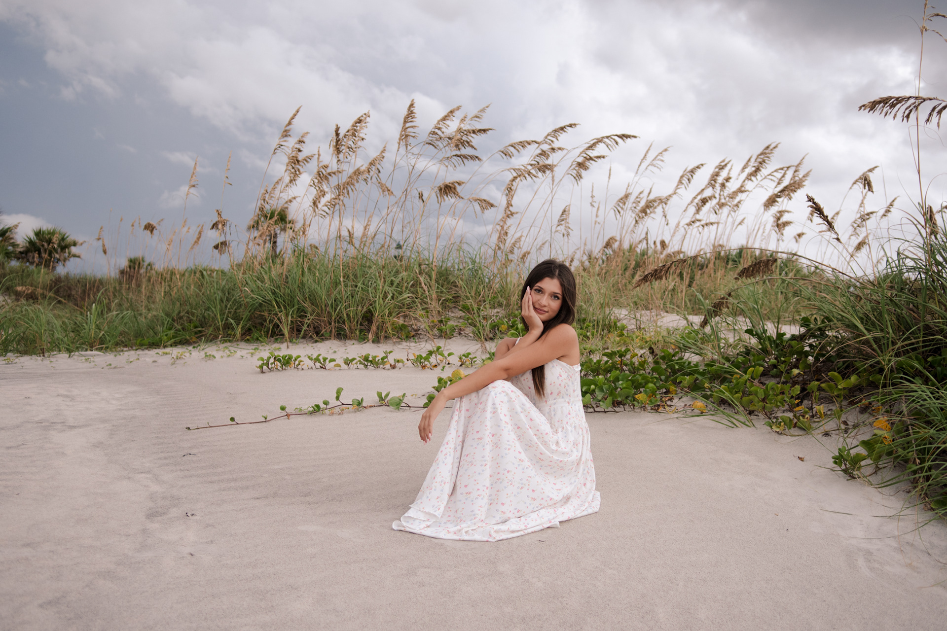 Senior photo in Ponce Inlet dunes, Mia sitting in a white floral dress surrounded by sea oats and sandy grasses.
