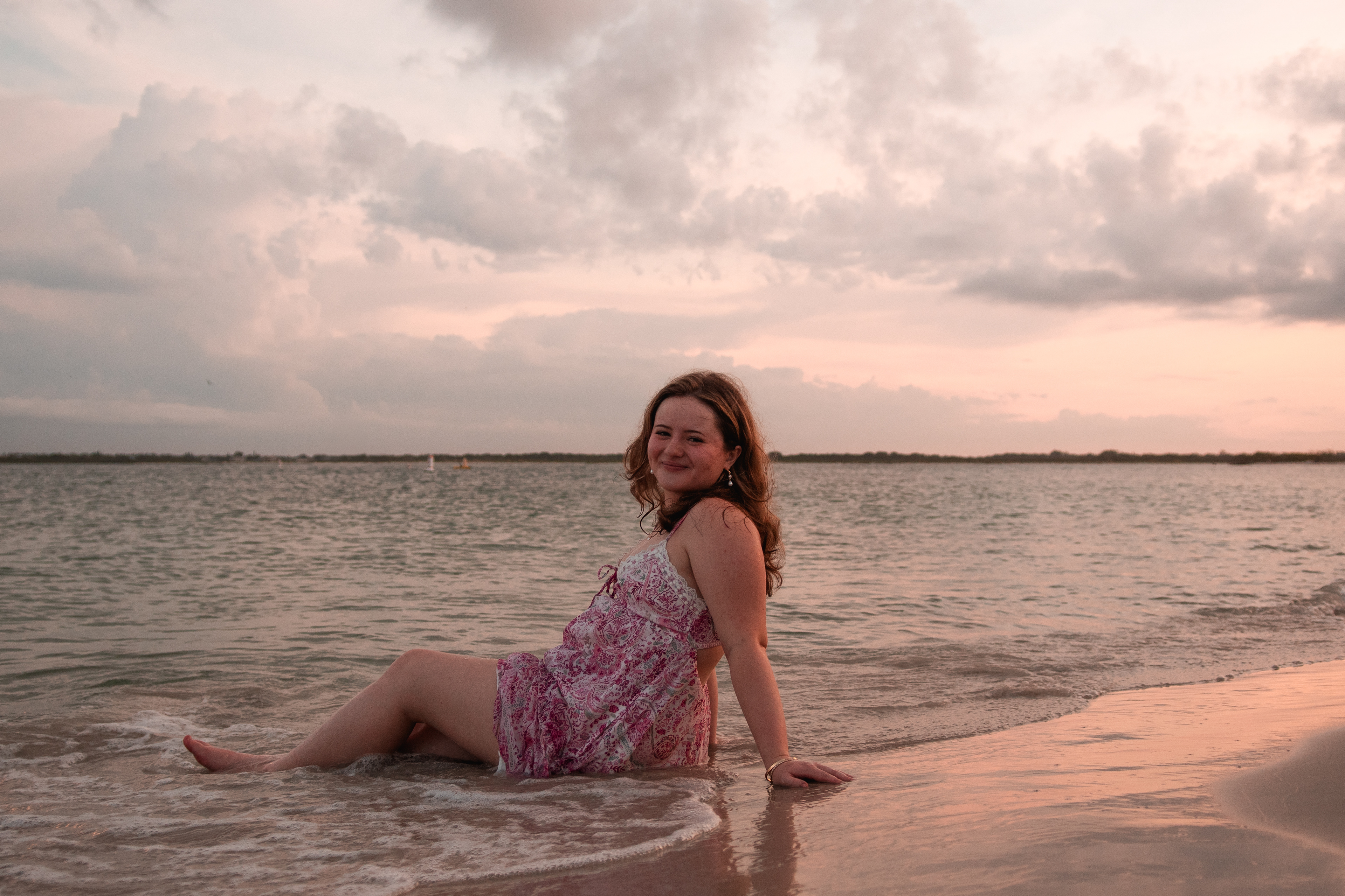 Daytona Beach area senior photos, Nelly smiling while seated at the water’s edge in pink dress, ocean glow behind her.