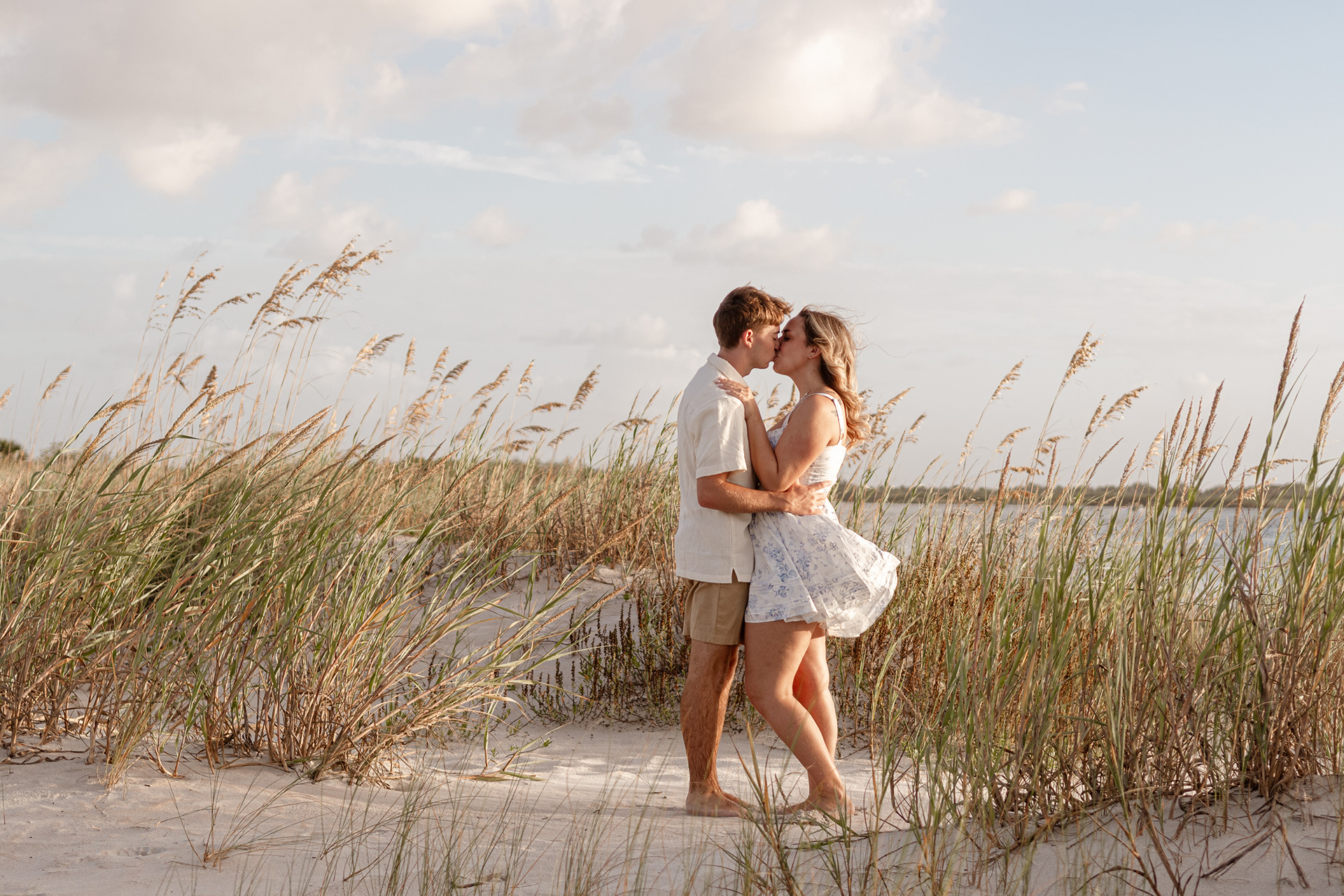Engagement photos in New Smyrna Beach dunes, couple embracing among sea oats and soft light.