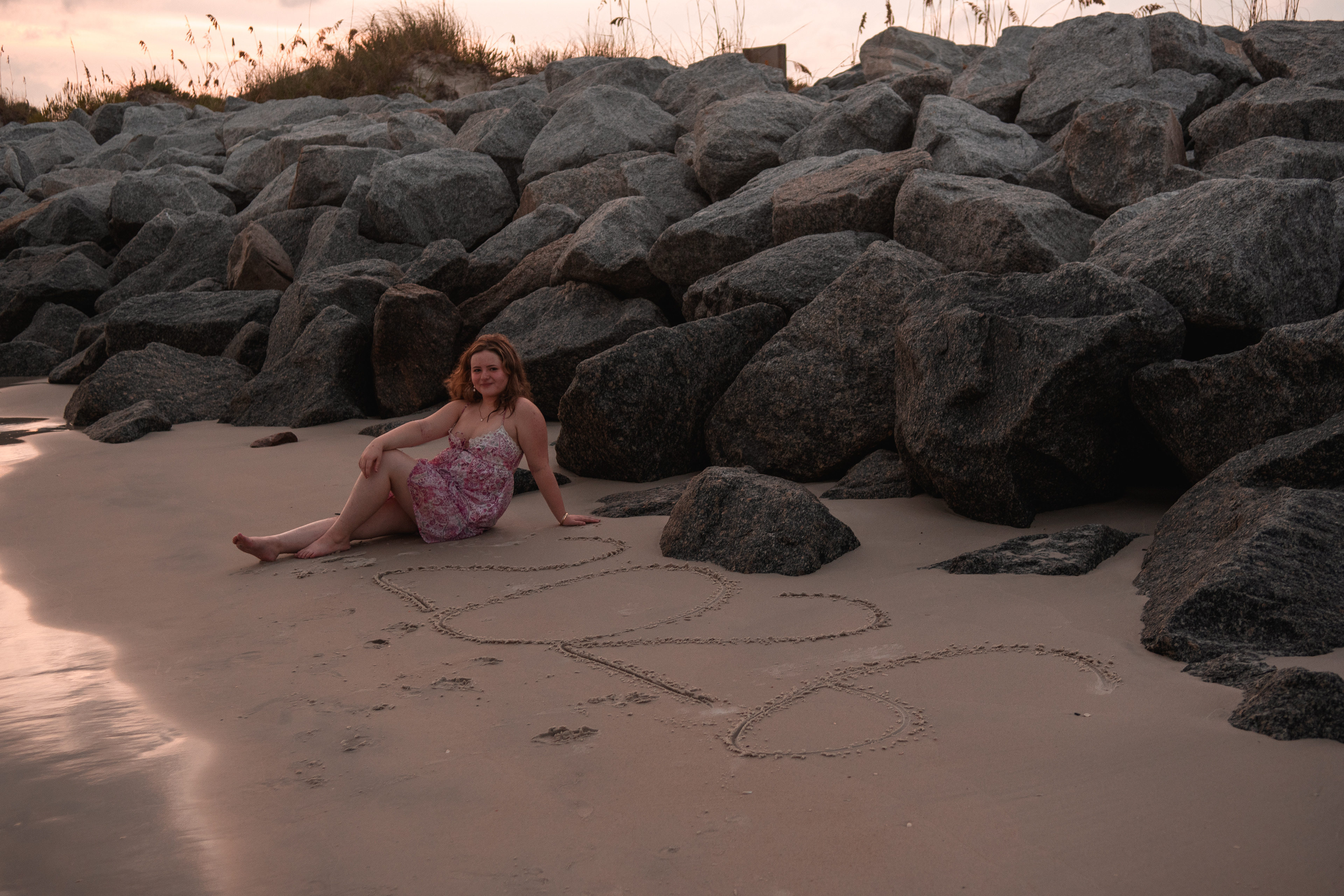 Senior photo in Ponce Inlet FL, Nelly sitting by large rocks on the shoreline, pink floral dress at golden hour.