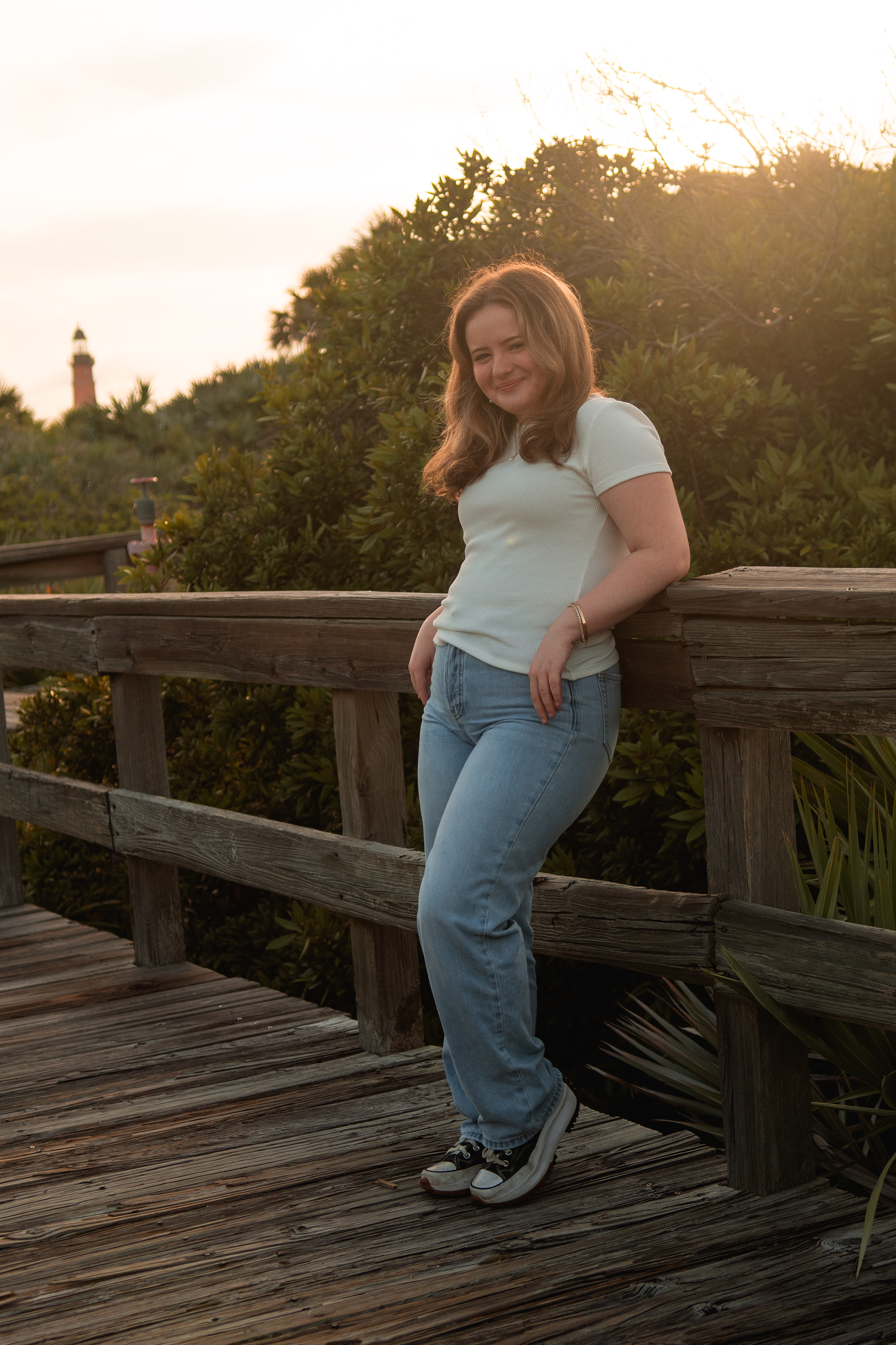 Senior portraits at Ponce Inlet, Nelly leaning against the boardwalk railing in a white shirt and jeans during golden hour.