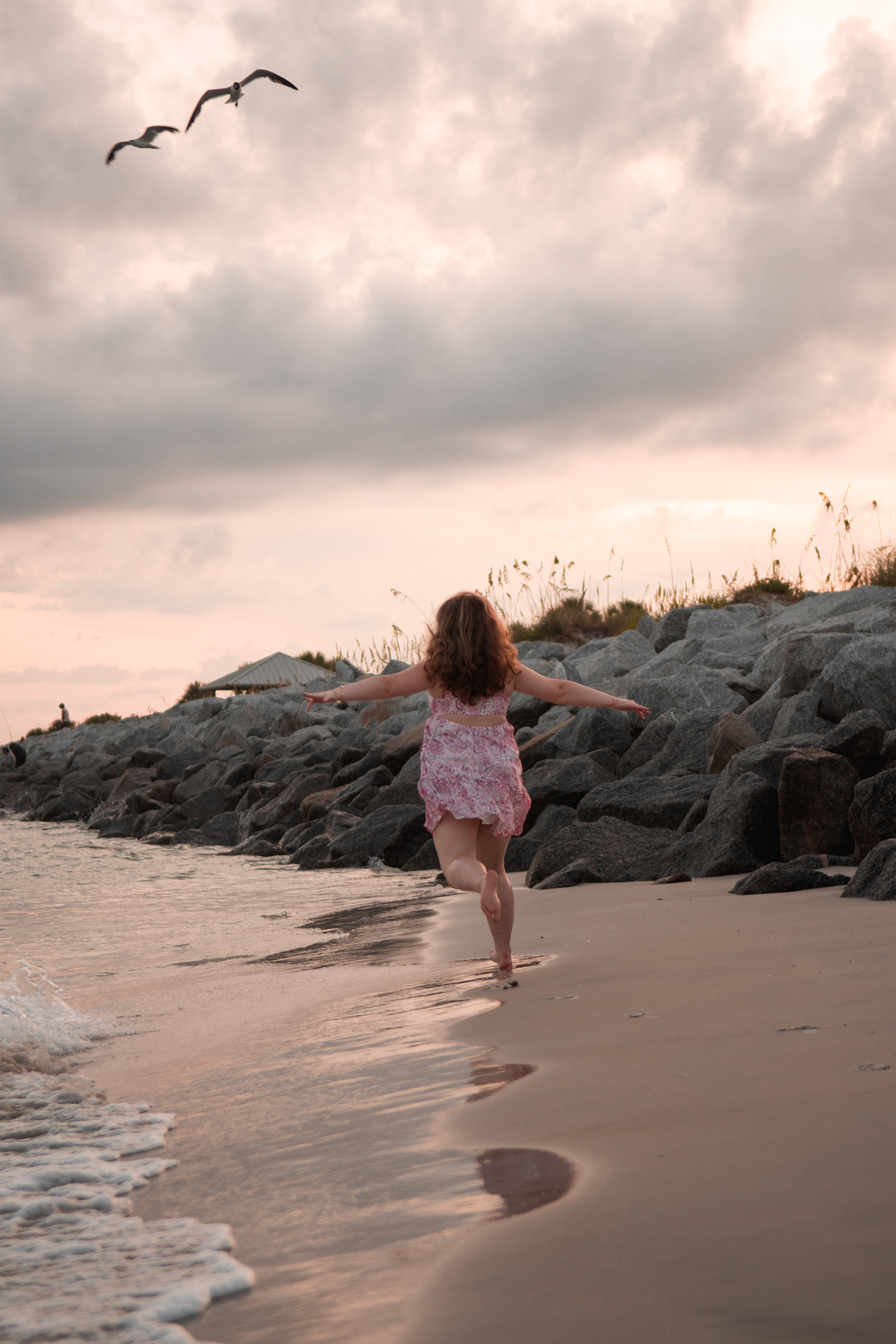 Senior portraits at Ponce Inlet FL, Nelly walking toward the ocean waves in a pink floral dress with birds flying overhead.