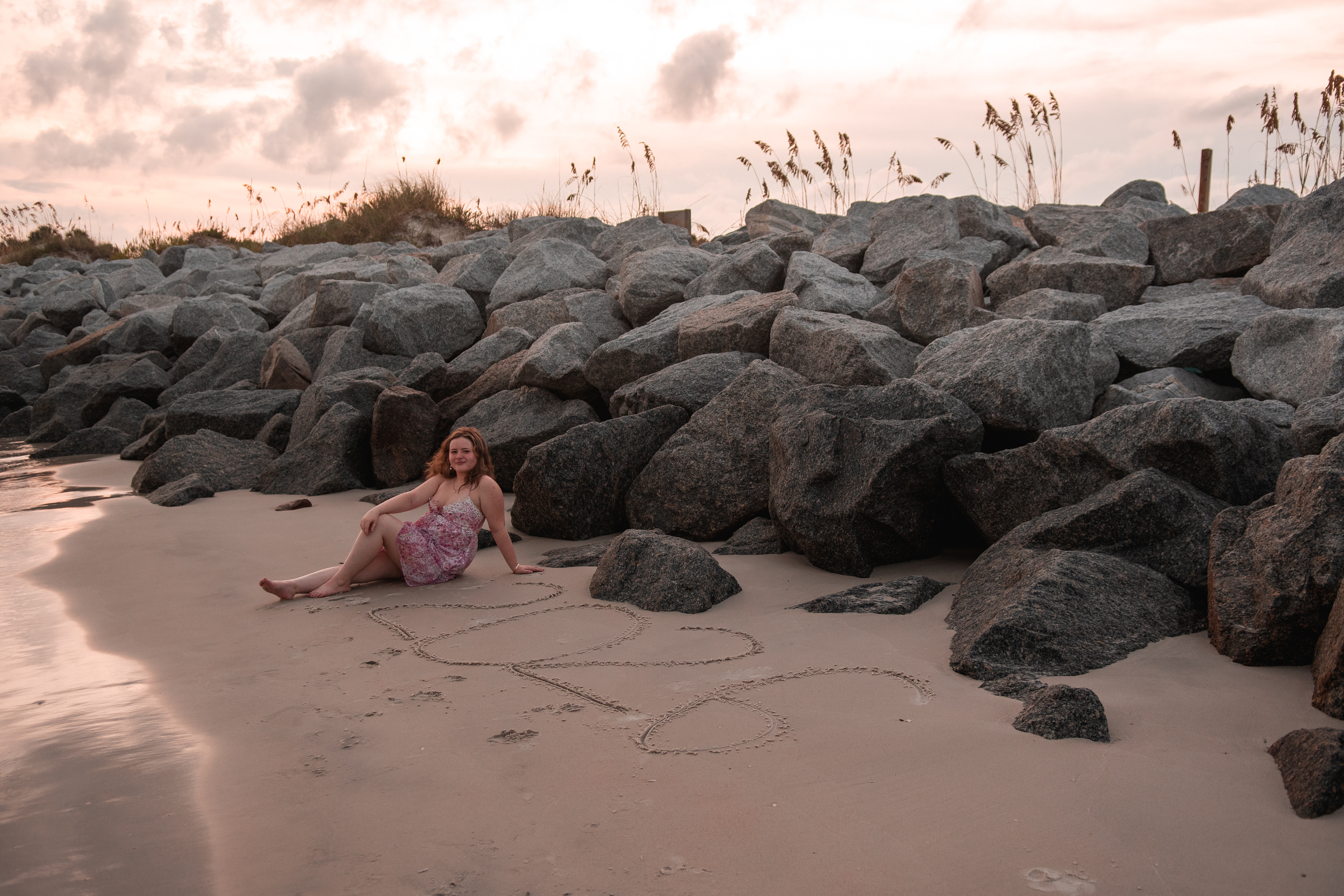 Daytona Beach senior portraits, Nelly sitting near inlet rocks with “2025” written in the sand.