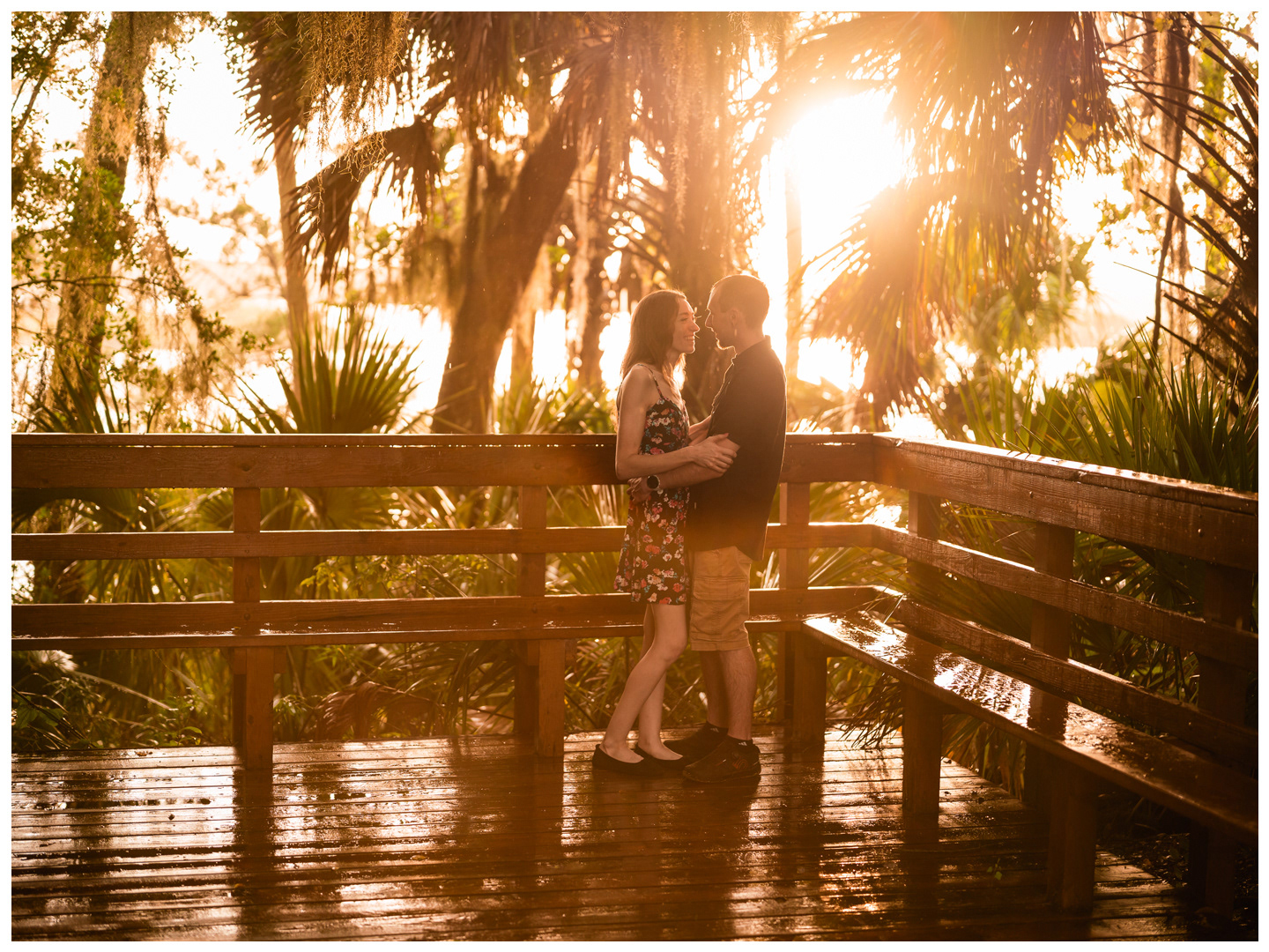 Wide portrait of a couple embracing on a wooden bridge beneath sunlit trees at Washington Oaks.