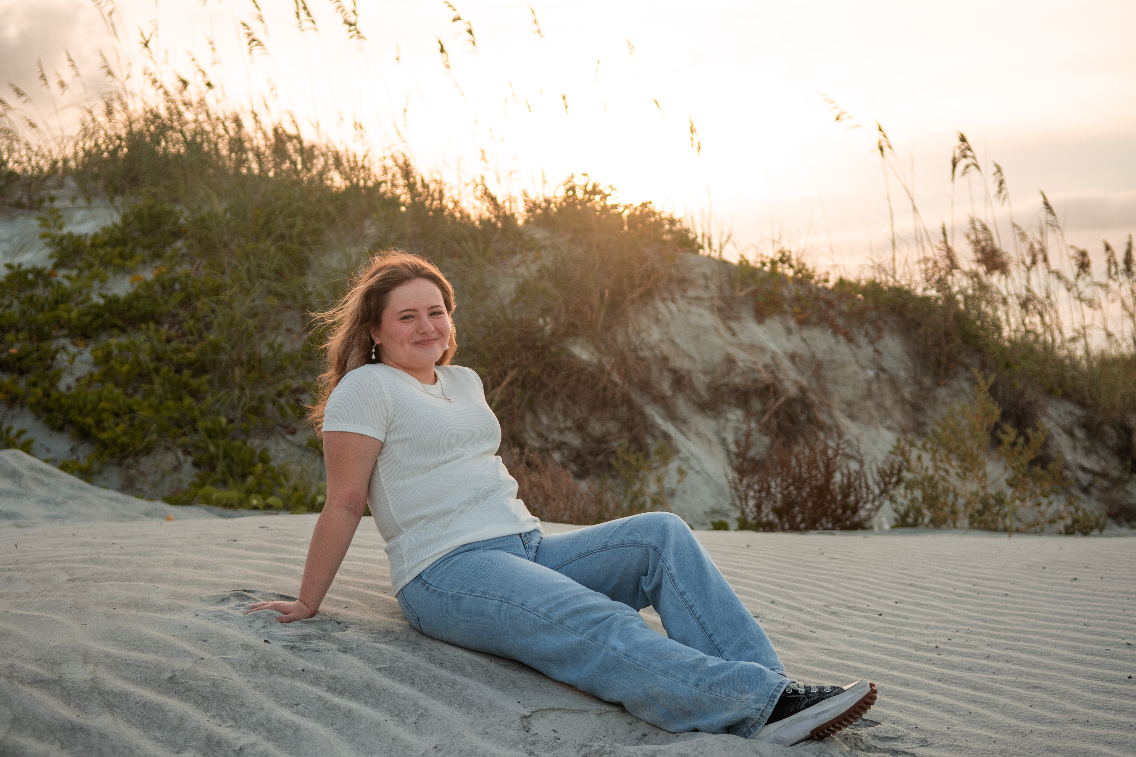 Daytona Beach senior photos, Nelly sitting on sand dunes in jeans and white top with sea oats and sunset glow.