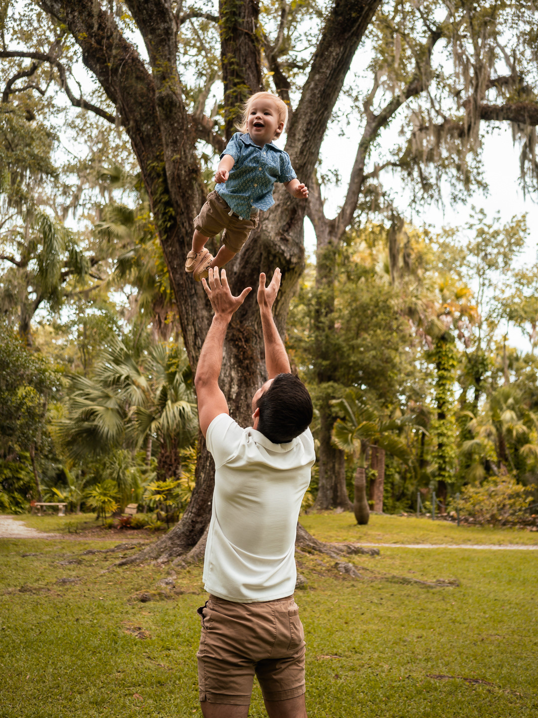 Dad lifting his youngest son high into the air beneath tall trees at Dunlawton Sugar Mill Gardens.