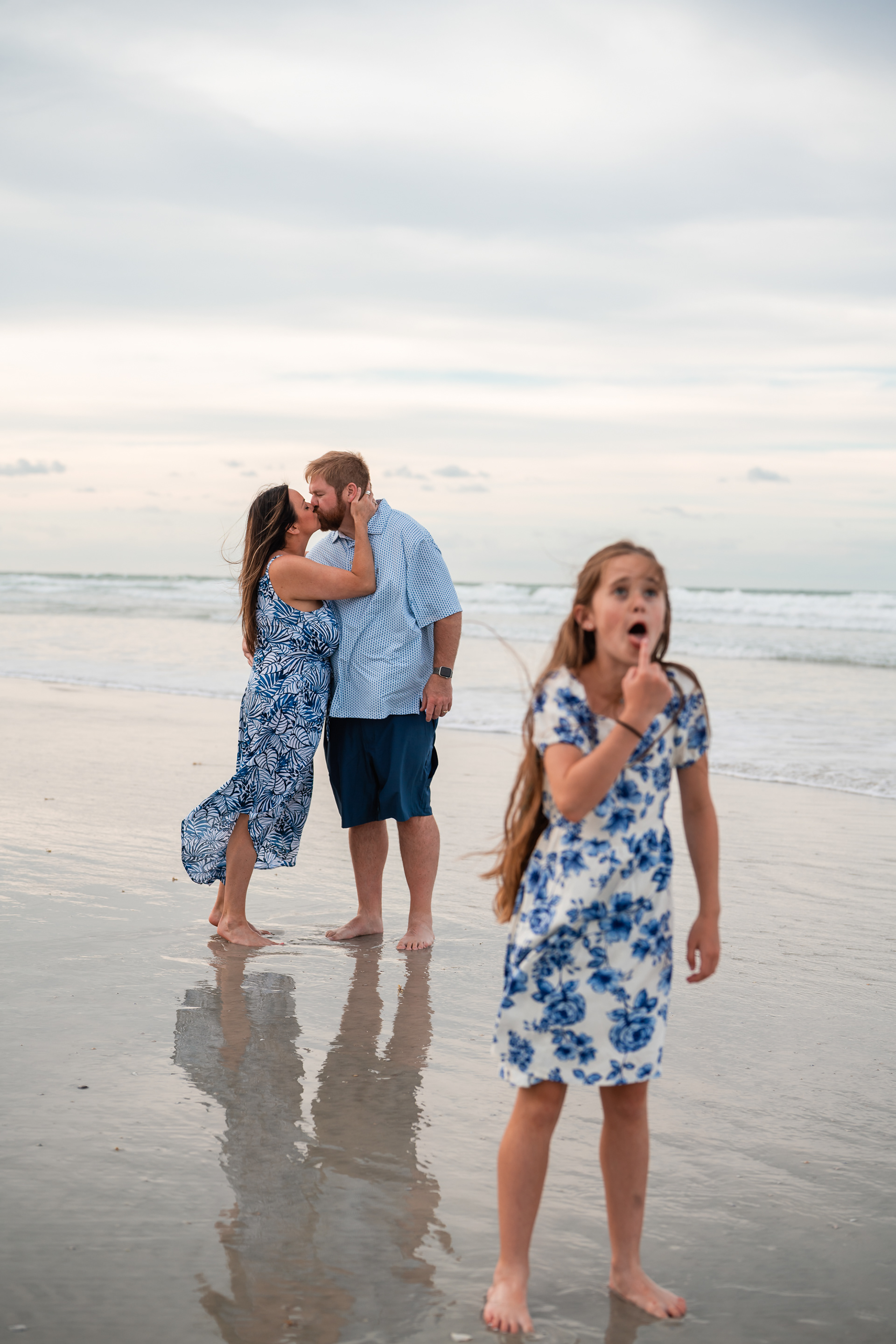 Couple kissing on the beach with their child in the foreground during a family photo session.