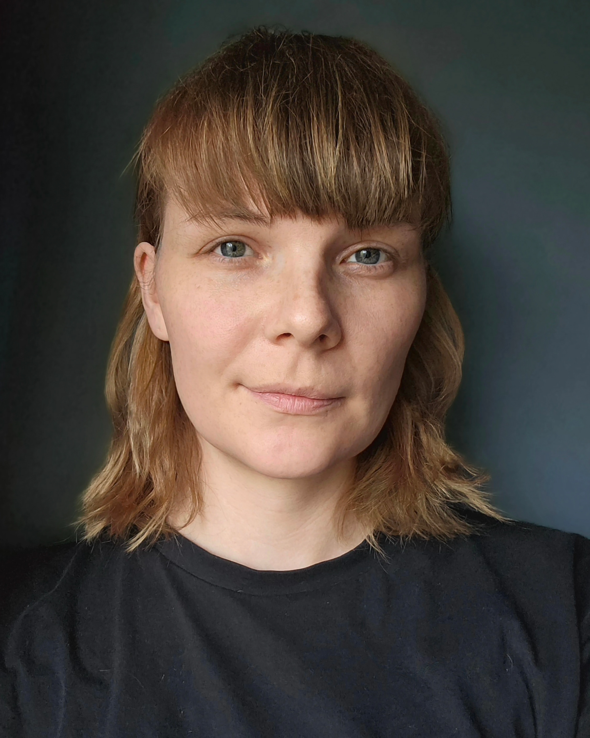 emelie is dark blond, has freckles and smiles, in front of a dark background