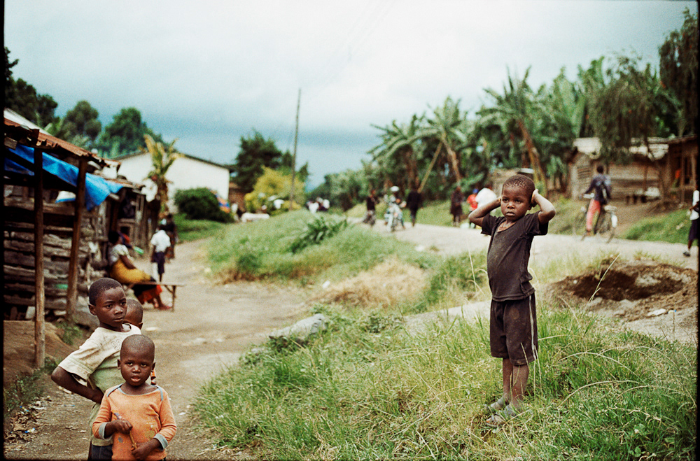 Two young boys watch the Mzungu (white person) curiously in the main hub of the village. Locals can be seen walking along the main road, going about their daily business