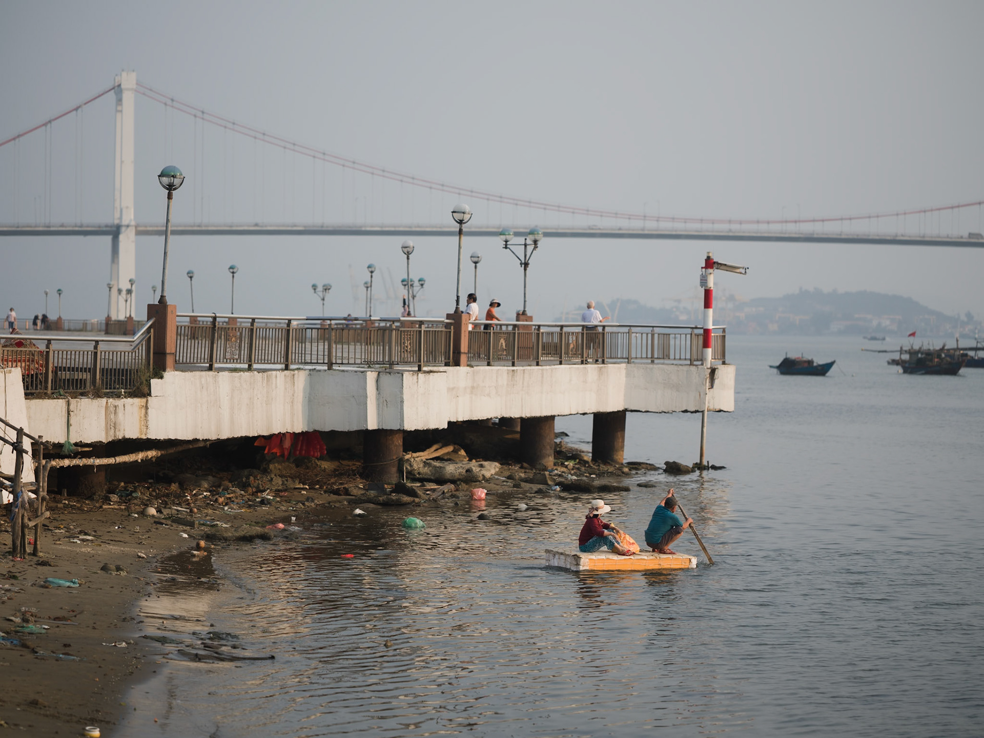 Two people paddling a small raft toward a fishing boat on the Hàn River in Đà Nẵng, Apr. 2026 Part of the VN26 Color series