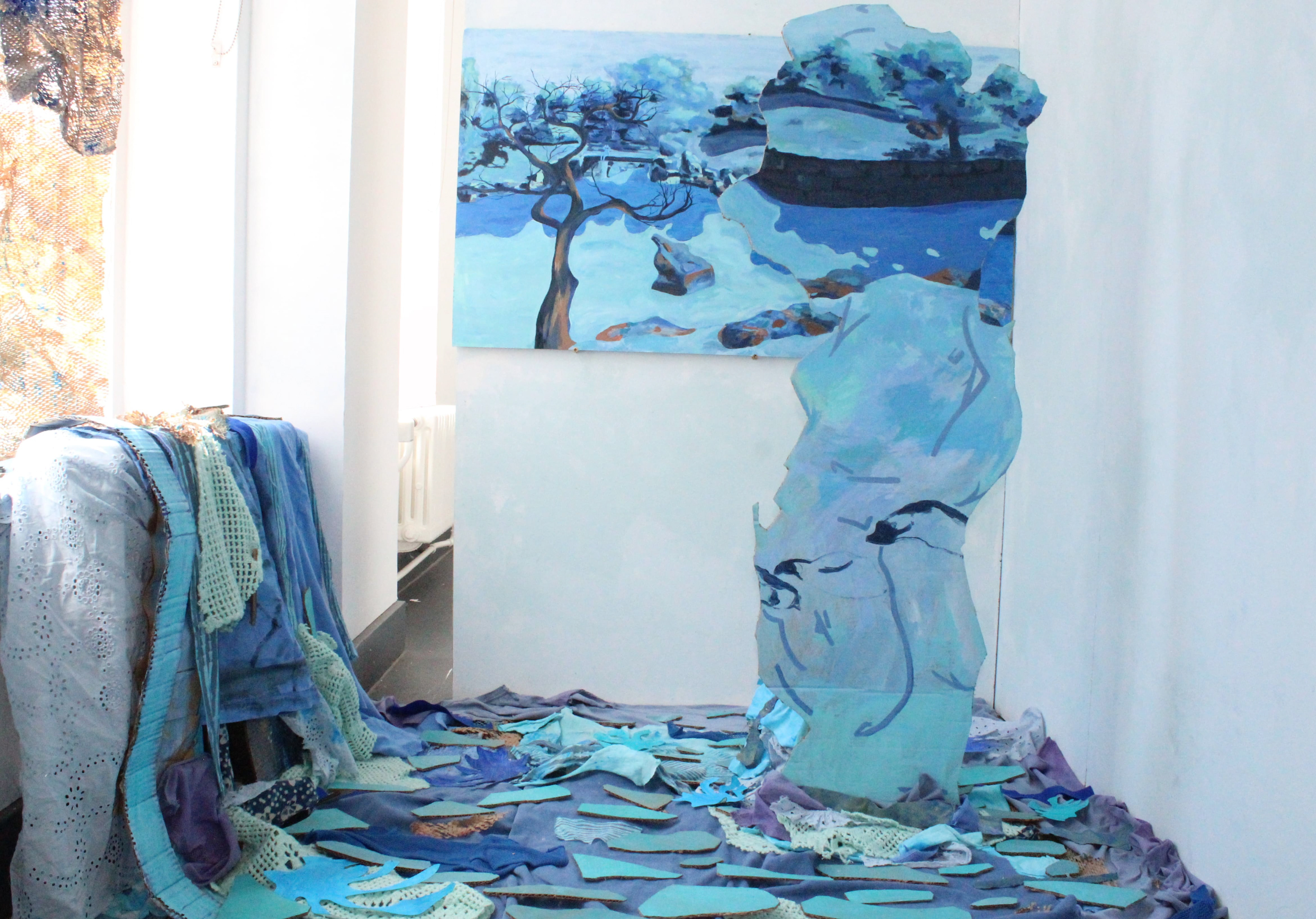 “Drifting Movements” Fia Cavozzi, Mixed fabrics, acrylic on wood, paper and cardboard, 2026 “Drifting Movements: I wanted a main painting that draws you into the work. This installation moves when the viewer changes their viewing angle as you move around the installation. The painting perspective shifts due to the cardboard cutout. This colour scene extends across the space, with different tones of blue, light greens and oranges to complement the fabric and cardboard to create its own separate space.  In the landscape painting, a cat sits on the bridge. This cat is a figment of my imagination and just exists in the world, exploring boundaries within the world. The cat is also in the fabric pool below, blending blue into water. The illusion of dissolving water gives the impression that you are closer than you feel. You are not near, but you are close.” Instagram: @nothingsquid_ 