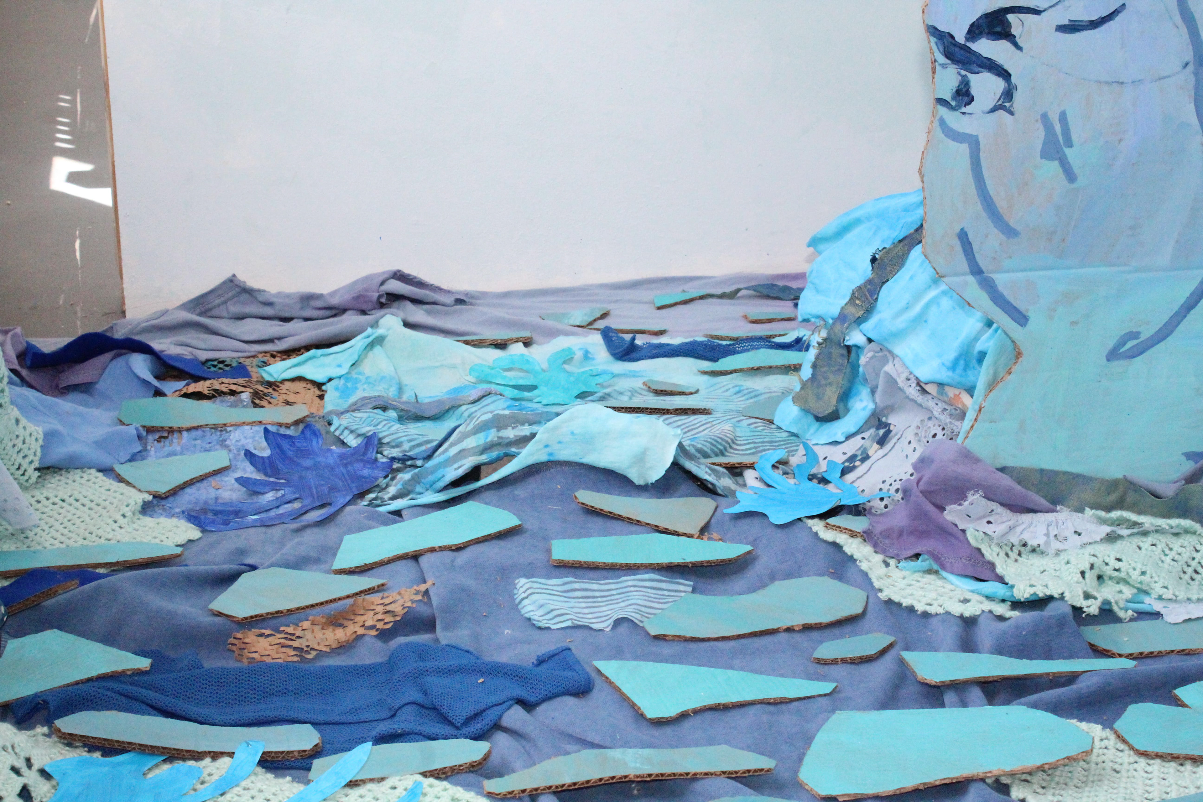 “Drifting Movements” Fia Cavozzi, Mixed fabrics, acrylic on wood, paper and cardboard, 2026 “Drifting Movements: I wanted a main painting that draws you into the work. This installation moves when the viewer changes their viewing angle as you move around the installation. The painting perspective shifts due to the cardboard cutout. This colour scene extends across the space, with different tones of blue, light greens and oranges to complement the fabric and cardboard to create its own separate space.  In the landscape painting, a cat sits on the bridge. This cat is a figment of my imagination and just exists in the world, exploring boundaries within the world. The cat is also in the fabric pool below, blending blue into water. The illusion of dissolving water gives the impression that you are closer than you feel. You are not near, but you are close.” Instagram: @nothingsquid_ 