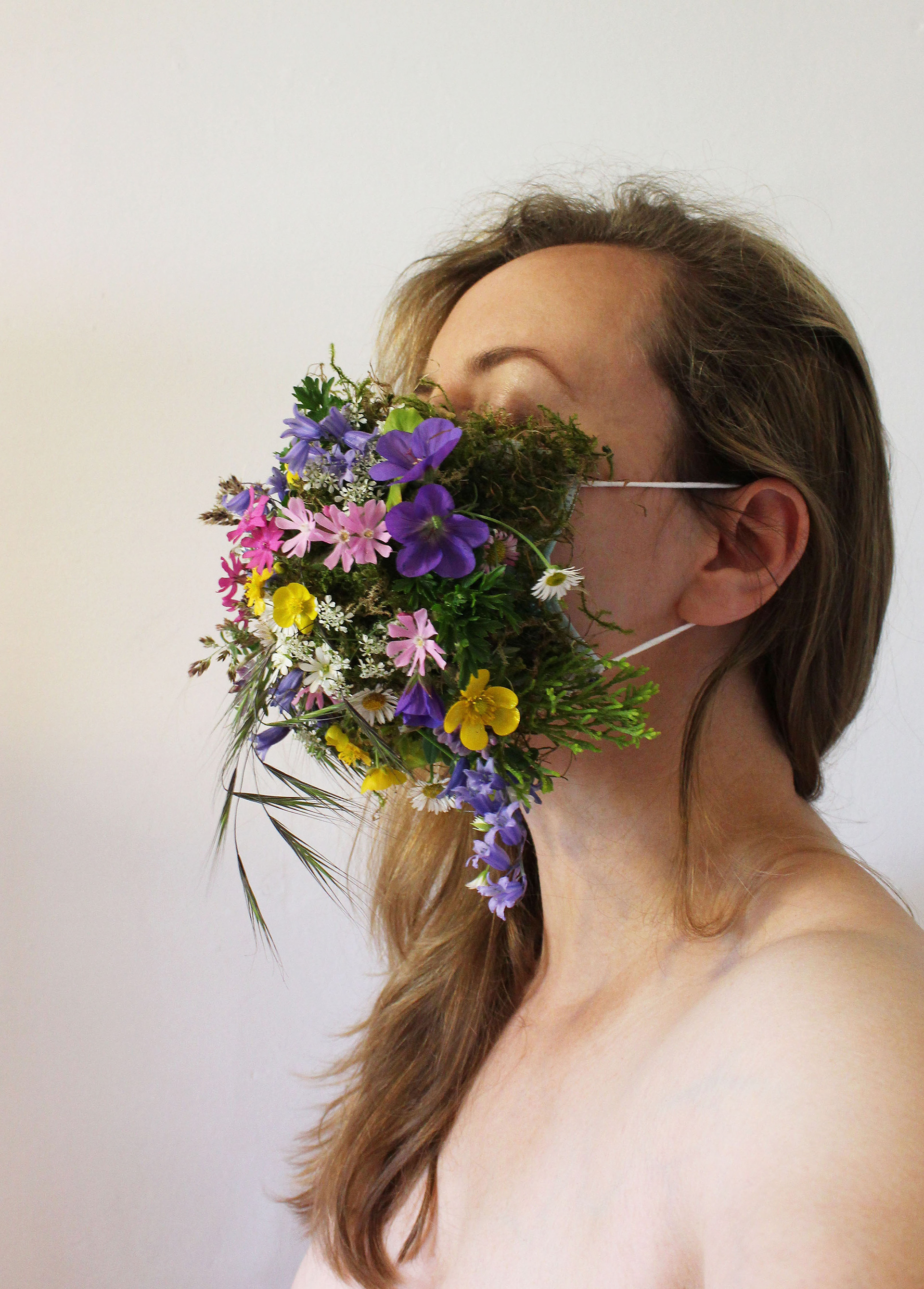 ‘’Rainbow Meadow’’ Estelle Woolley, Photography and wildflowers, 2020 ‘’Through her practice Estelle Woolley often explores ways that we work with or against nature; how we react and intervene, and how nature responds back at a domestic level and beyond. Through a minimal and poetic use of materials, which are often very fragile, she questions possible narratives and layers of meaning. By subtle manipulation, she hopes to renew a sense of curiosity, while always highlighting the beauty inherent in the forms used. Over lockdown, she has produced a series of photographic self portraits wearing facemasks composed of delicate and ephemeral natural materials. These have been collected from her daily walks, where she has been homing in on her immediate surroundings, paying close attention to the plant life as it comes in and out of season. Spending lockdown in the Cheshire countryside on her family’s farm, Estelle has been immersed in the changing seasons. Rainbow Meadow was created in late Spring, when many different coloured wild flowers and grasses began to emerge in the hedgerows, such as pink campions, buttercups, bluebells and forget me nots. As one of her earlier masks in the series, Rainbow Meadow was made during a time when every Thursday evening the country went outside to clap and show their support for the NHS. The rainbow became a symbol of this support, which inspired the thinking behind the mixed colour palette. Other masks in the series have been made from materials such as dandelion clocks, nettles, thistles, lavender, wild chamomile, honeysuckle, chive flowers, oak leaves, robin’s pincushions, forget me nots, clover leaves, moss, sycamore leaves, foxgloves, bees, butterflies, and even a wasp nest! Each with their individual narratives.’’
