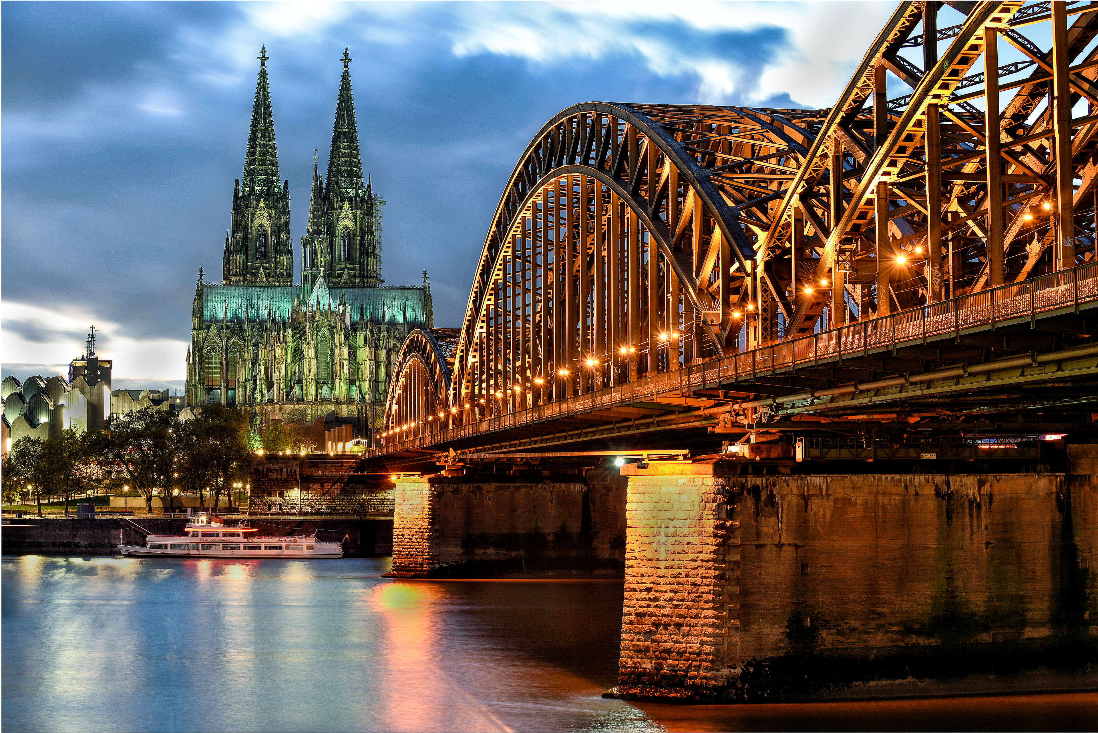 A low-angle view of the Hohenzollern Bridge's massive steel arches leading to the illuminated Cologne Cathedral at night