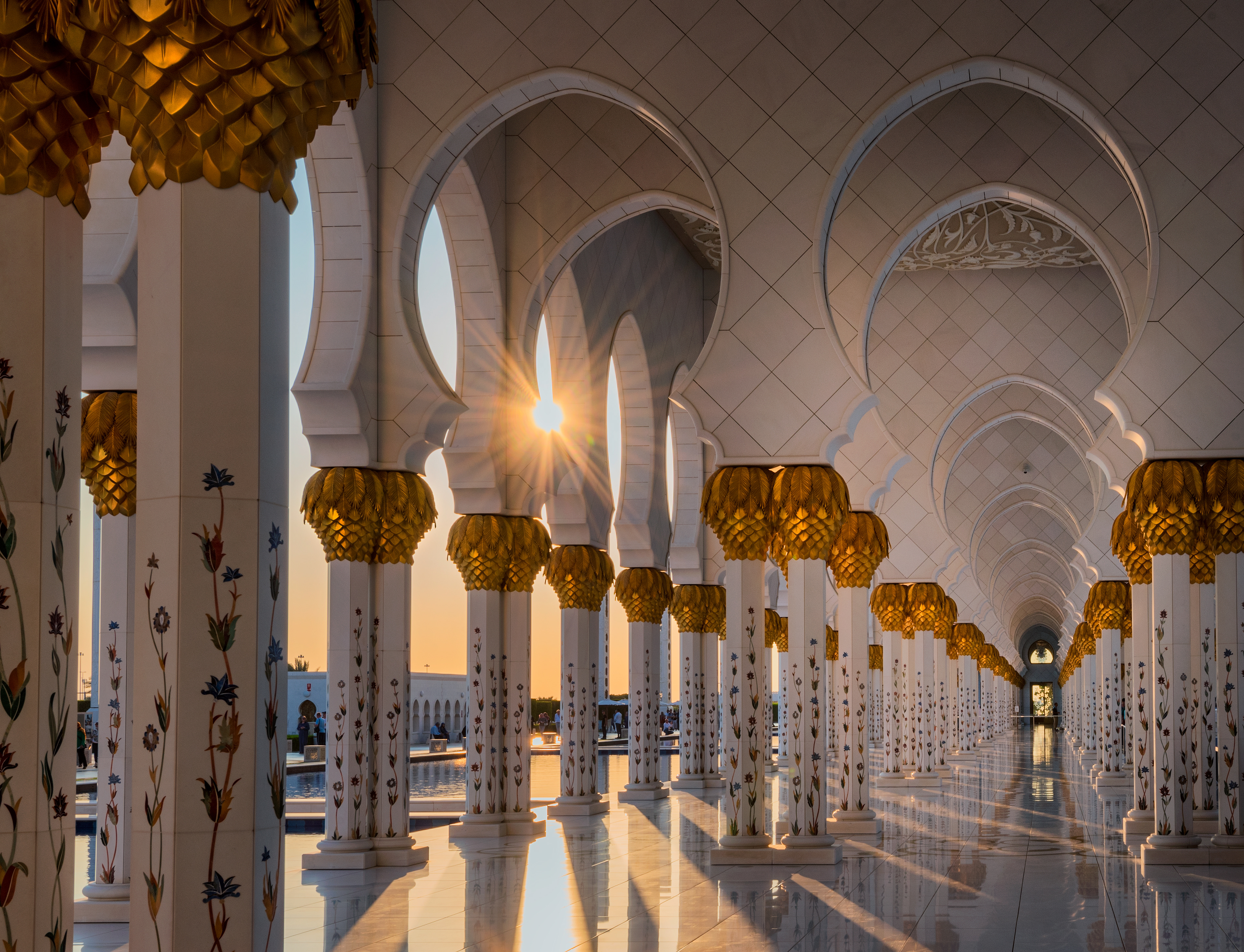 Sunlight bursts through the floral marble pillars of the Sheikh Zayed Grand Mosque, casting long shadows across the reflective courtyard