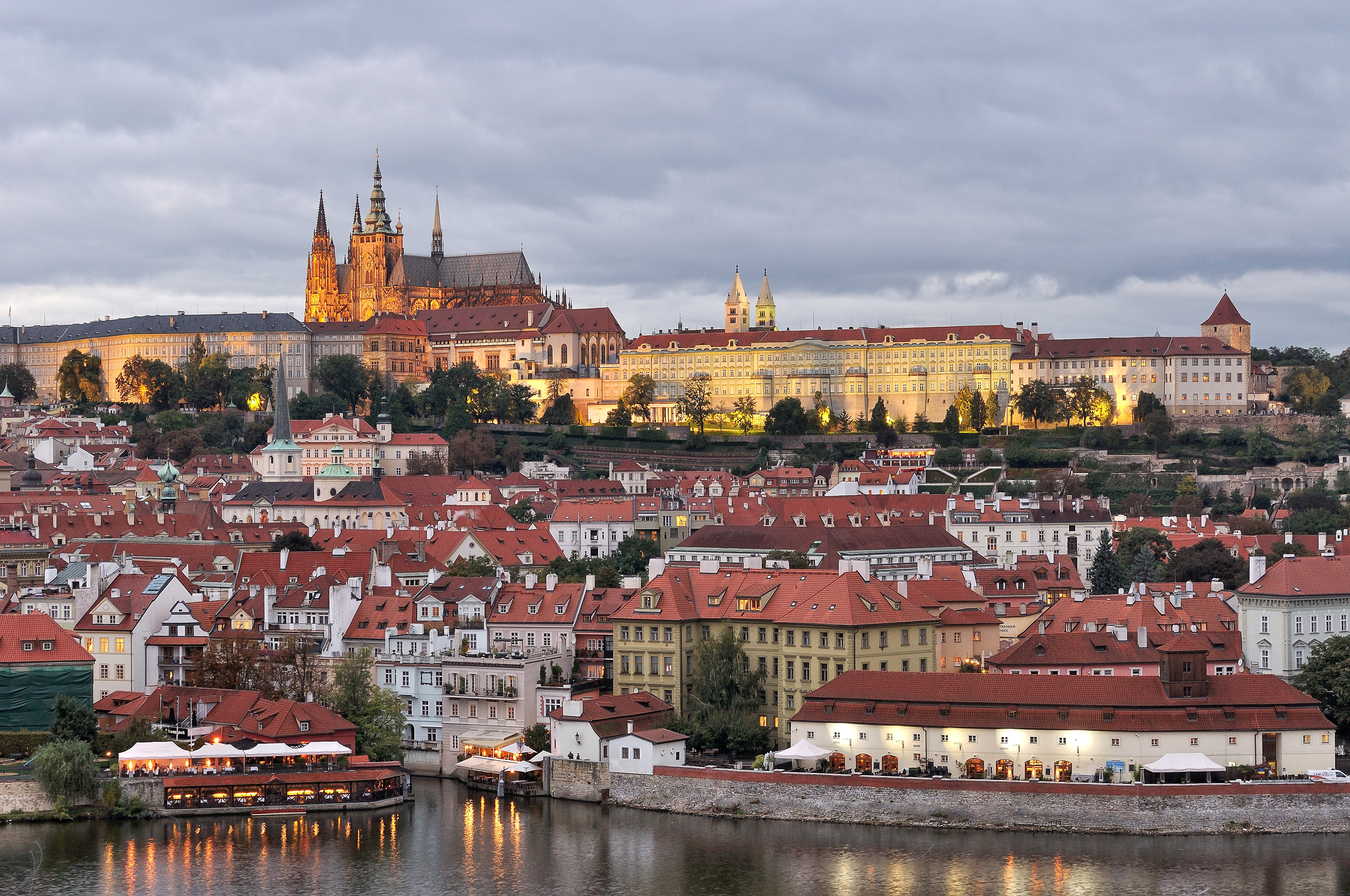 A panoramic evening view of Prague, featuring the illuminated Prague Castle and St. Vitus Cathedral rising majestically above the Vltava River