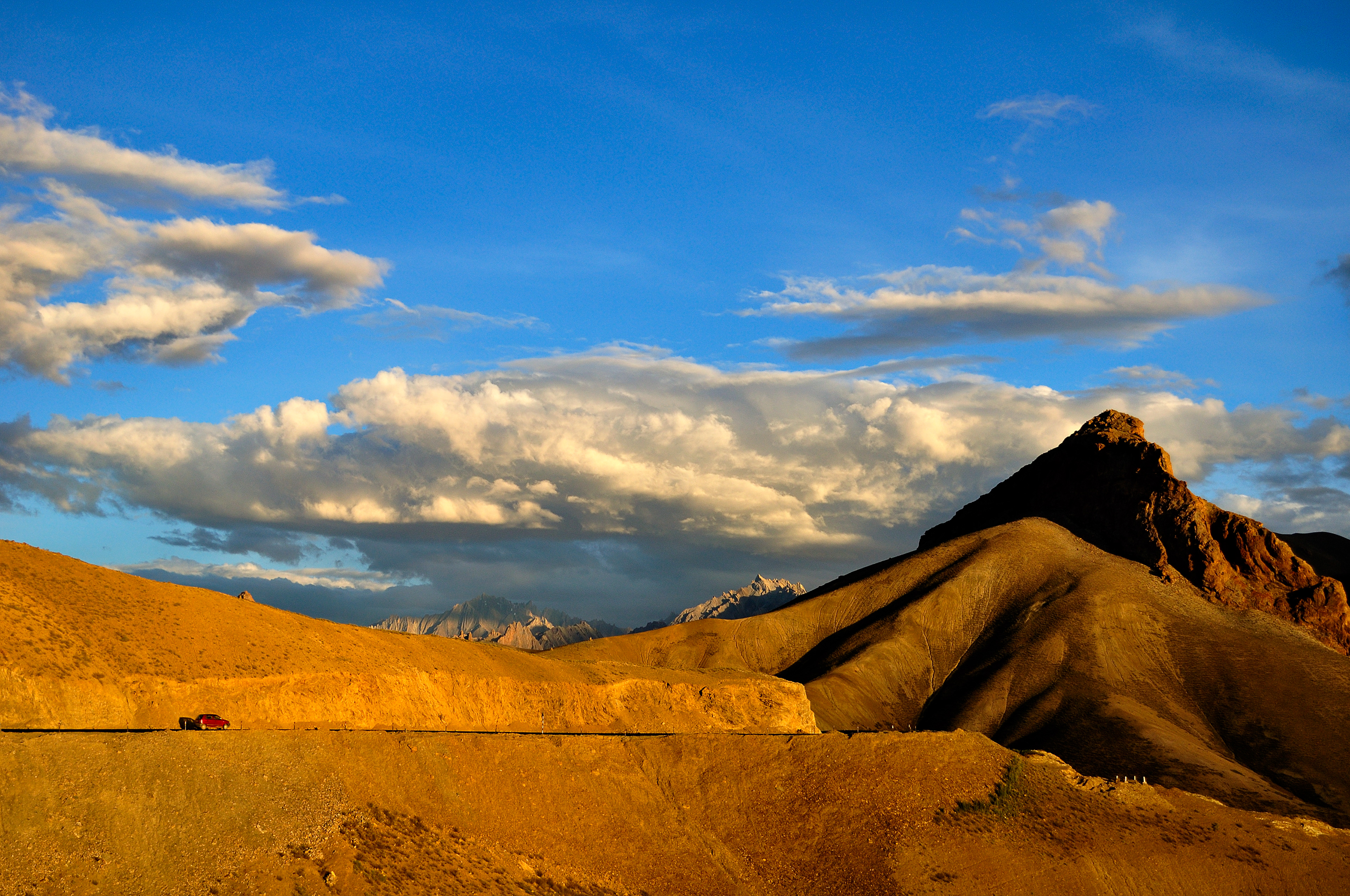 Golden hour sunlight illuminates the rugged, barren mountains of Ladakh, creating a striking contrast against the deep blue Himalayan sky